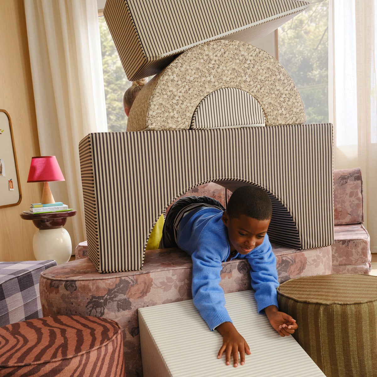 A smiling boy in a blue sweater crawls through a brown striped arch block, with a tan floral arch and striped piece stacked on top.