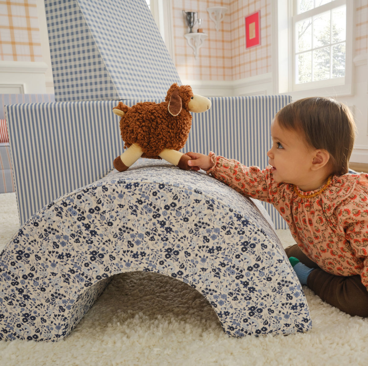 A toddler in a floral shirt kneels on a white shag rug, reaching toward a brown plush sheep toy placed on top of a blue-and-white floral fabric arch. In the background, a child plays with a blue-and-white striped pillow and a blue-and-white checkered tent in a bright room.
