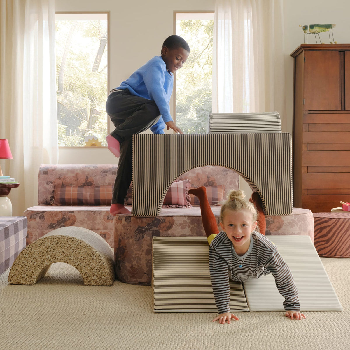 A boy climbs onto a brown striped arch block while a smiling girl crawls down two striped wedges, with a tan floral arch nearby.