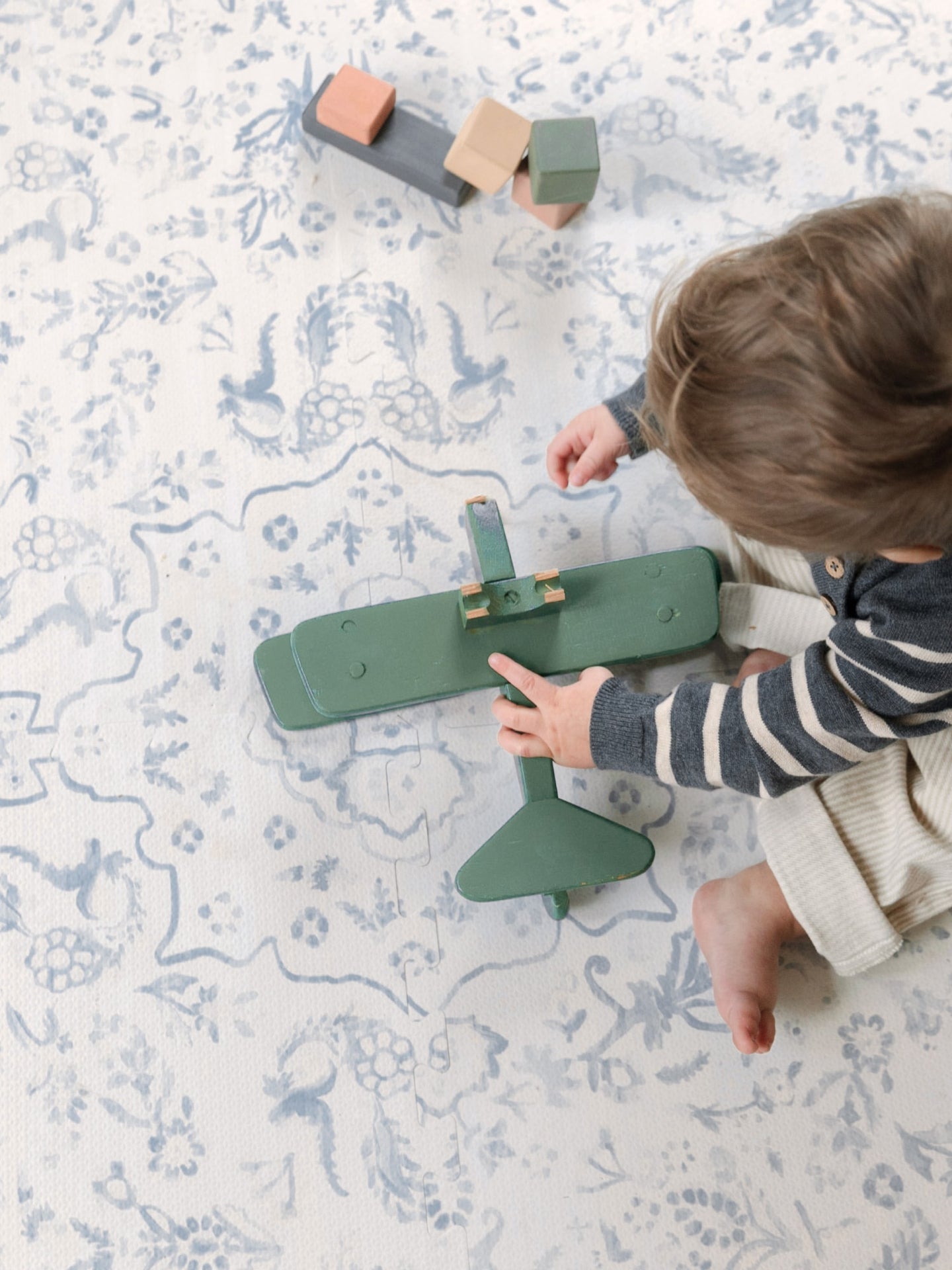 Child playing with a green toy airplane on a blue and white House of Noa Play Mat
