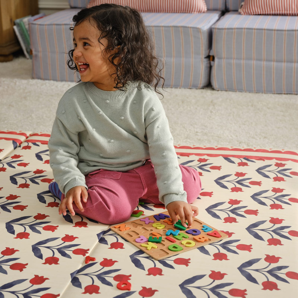 Smiling toddler sitting on the red tulip Delilah Carnation tumbling mat with a wooden alphabet puzzle