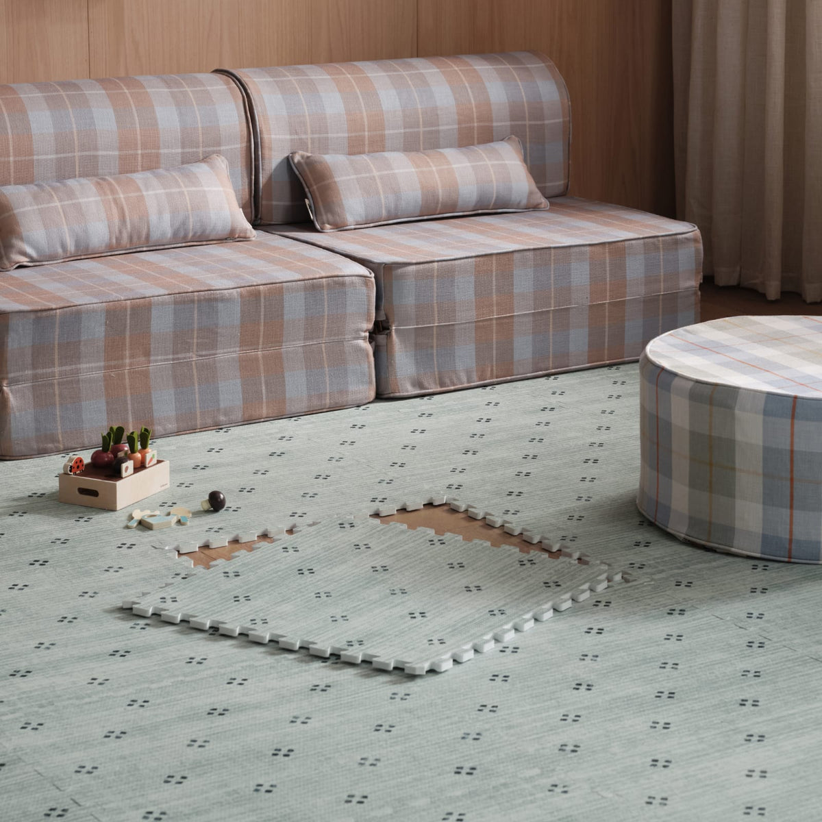 A seafoam Dover play mat covering a living room floor, with a few tiles lifted to show the wood floor beneath, next to a plaid modular sofa and round ottoman.