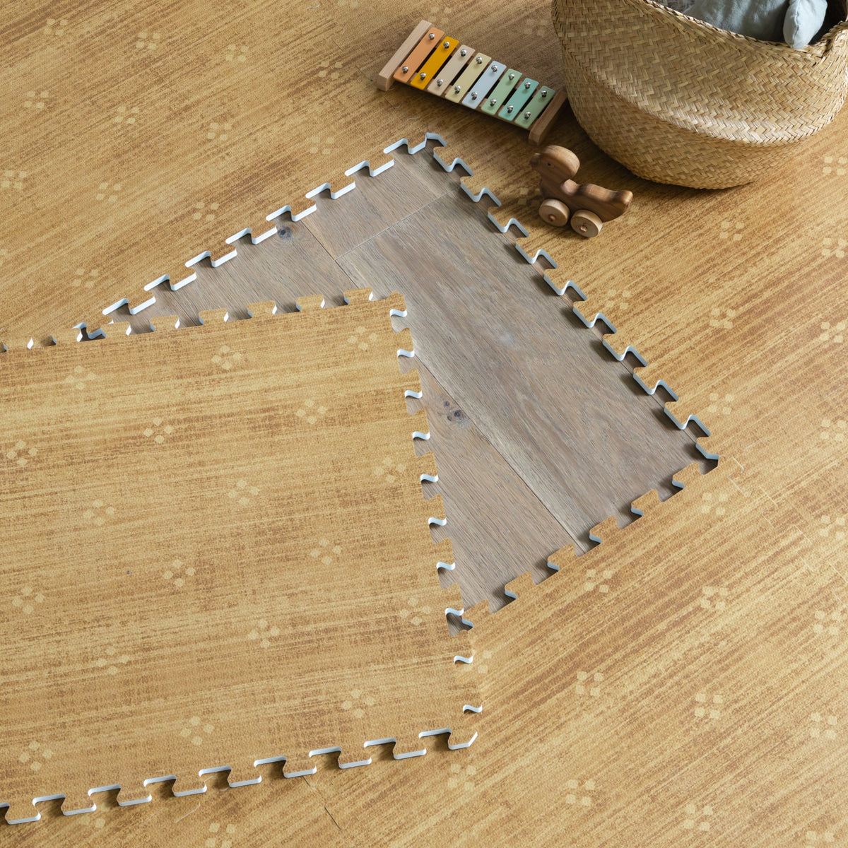 Two mustard foam play mat tiles lifted to reveal the floor beneath, with a woven basket and a wooden xylophone resting on the mat nearby.