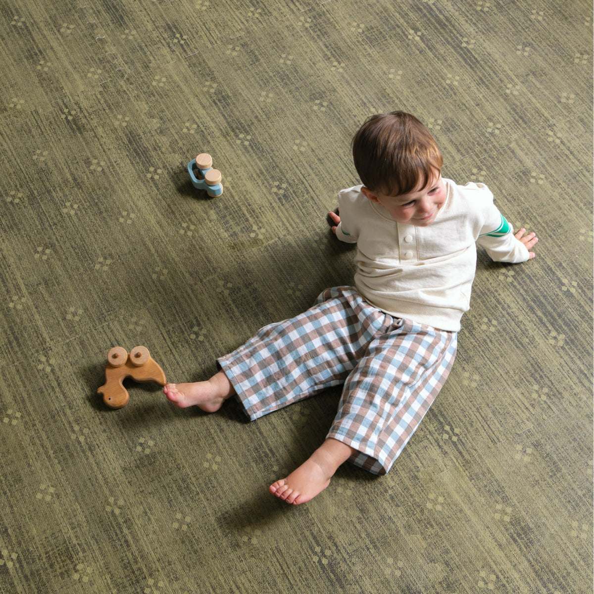 A toddler in plaid trousers sitting on an olive green foam play mat, seen from above, with a wooden pull toy beside them.