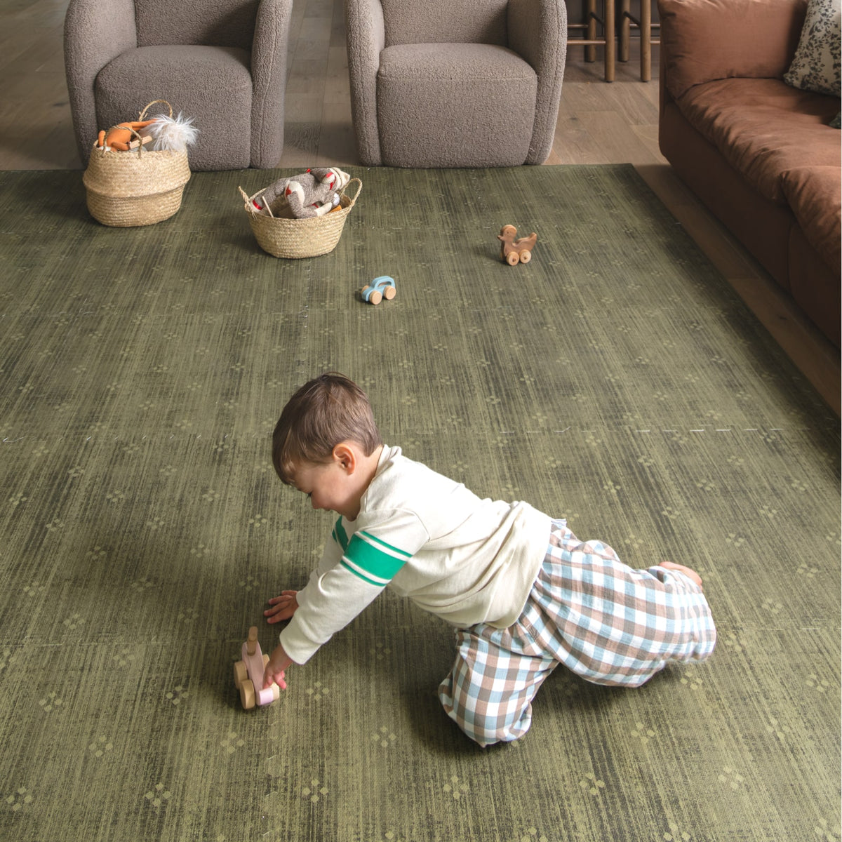 A toddler crawling toward a wooden toy on an olive green play mat in a living room with boucle chairs and woven baskets of toys.