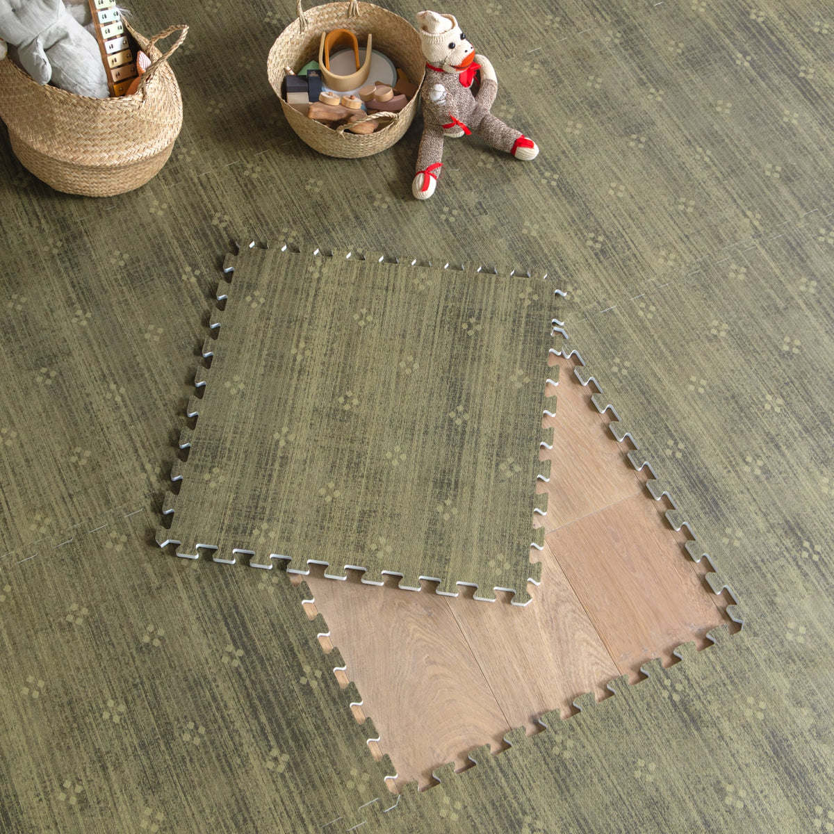 Olive green play mat tiles lifted at the corner to show the wood floor beneath, with woven baskets of wooden toys and a teddy bear nearby.