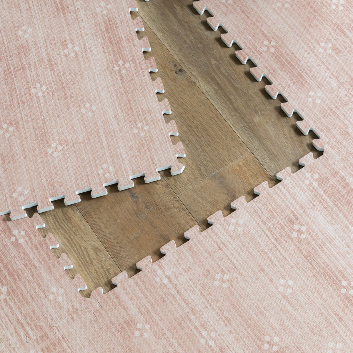 A close-up of dusty rose foam play mat tiles lifted at the corner to reveal the wood floor underneath, showing the interlocking puzzle edges.