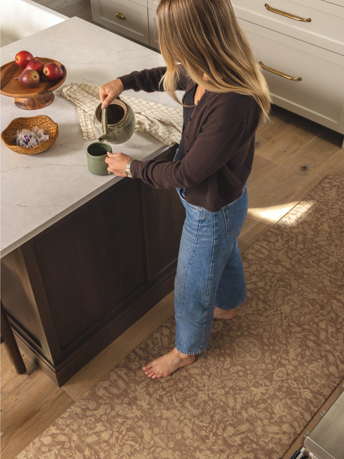A woman in a kitchen pours tea from a ceramic pot into a mug on a marble countertop. She is wearing a dark brown long-sleeve top and blue jeans, standing barefoot on a patterned brown area rug.