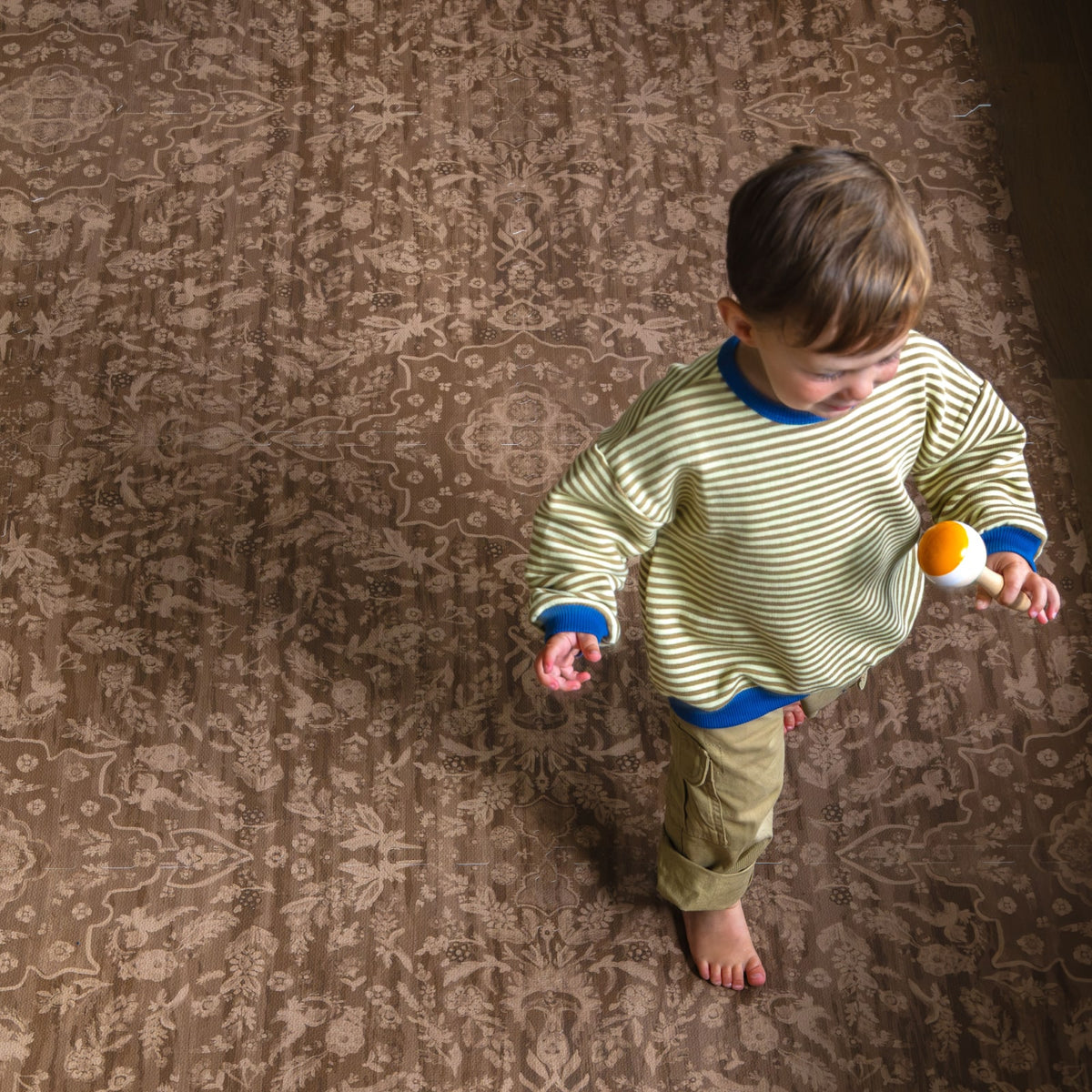 A young child in a striped sweater standing on a cocoa brown floral foam play mat, holding a wooden ball, seen from above.