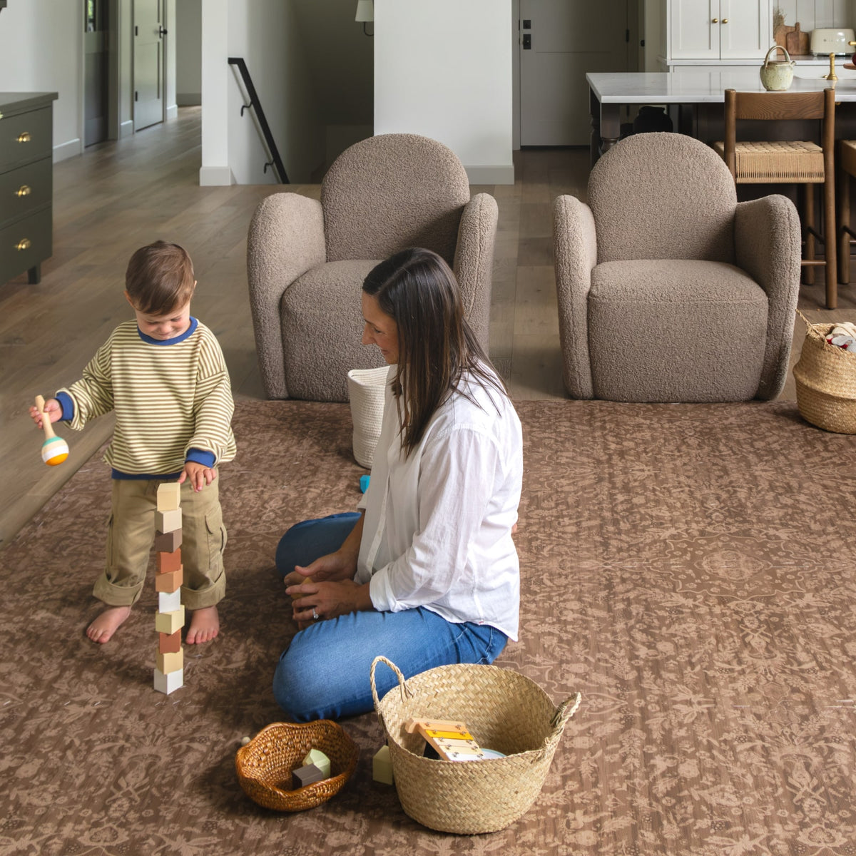 A parent and toddler stacking wooden blocks on a cocoa brown floral play mat in a living room with boucle chairs and woven toy baskets.