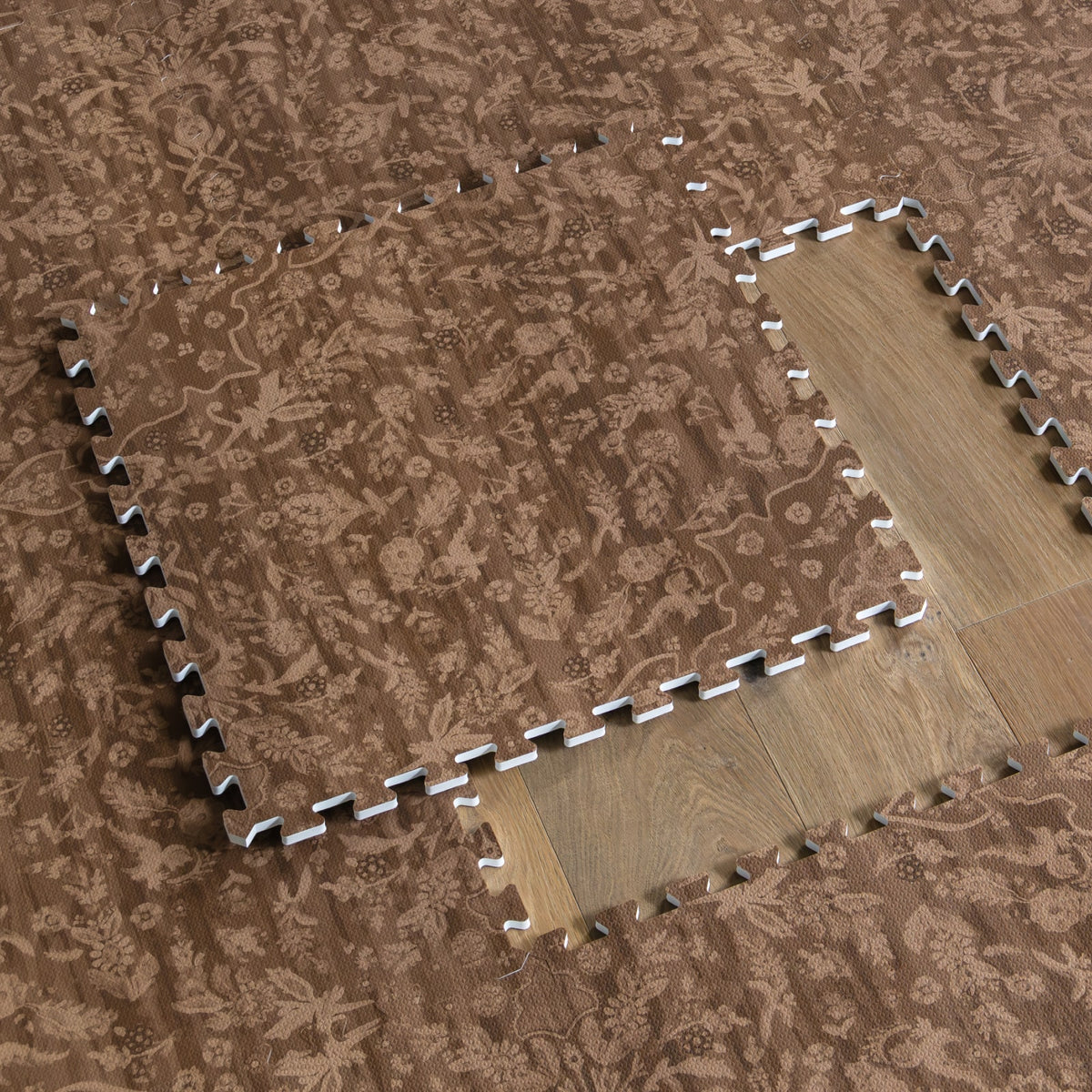 A cocoa brown floral play mat tile lifted to reveal the wood floor beneath, showing the interlocking puzzle edges of the foam mat.