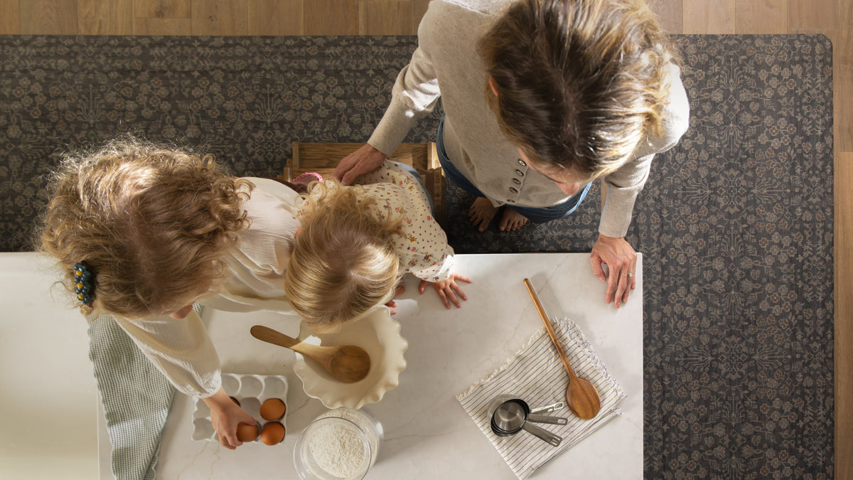 Overhead view of a woman and two young girls gathered around a white table, preparing food with eggs, flour, a wooden spoon, and measuring cups in a home kitchen setting.