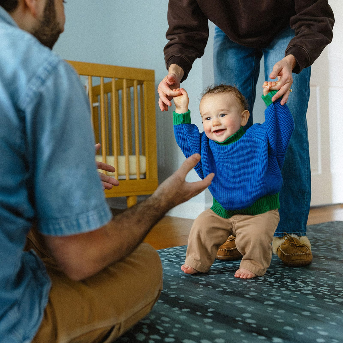 A smiling toddler in a blue sweater takes steps while holding a parent's hands on a deep turquoise Fawn play mat in a nursery.