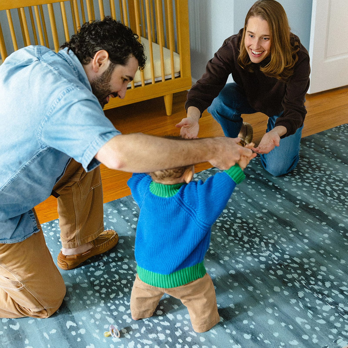 Two parents kneel on a deep turquoise Fawn play mat, cheering on a toddler learning to walk beside a wooden crib.