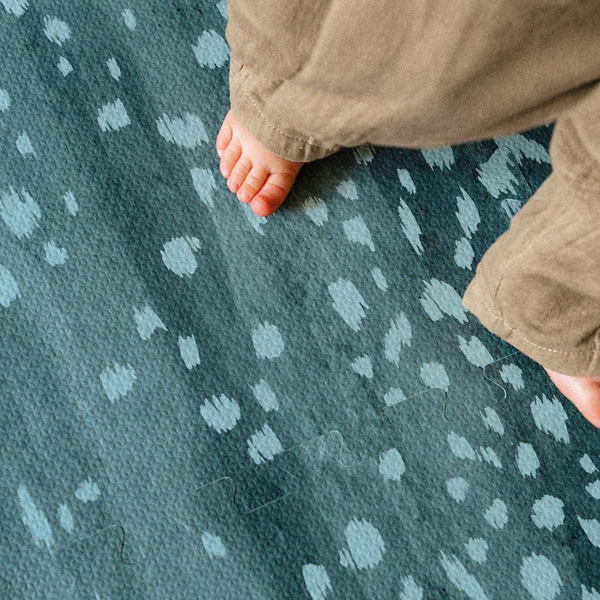 A close-up of a toddler's bare feet on a deep turquoise foam play mat with pale aqua fawn spots.