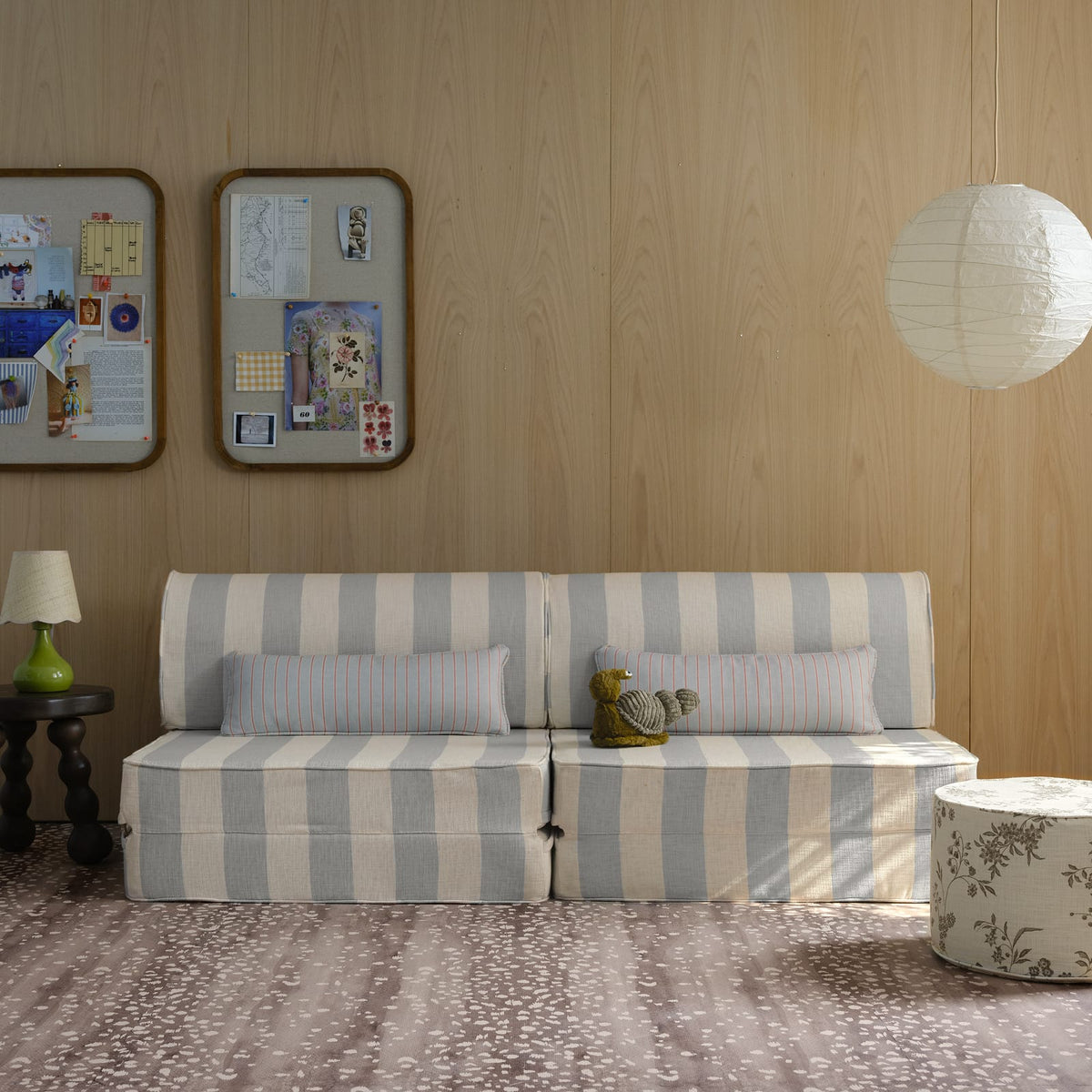 A walnut brown Fawn play mat covering the floor of a wood-paneled playroom with a striped modular sofa, a floral ottoman and a paper lantern overhead.