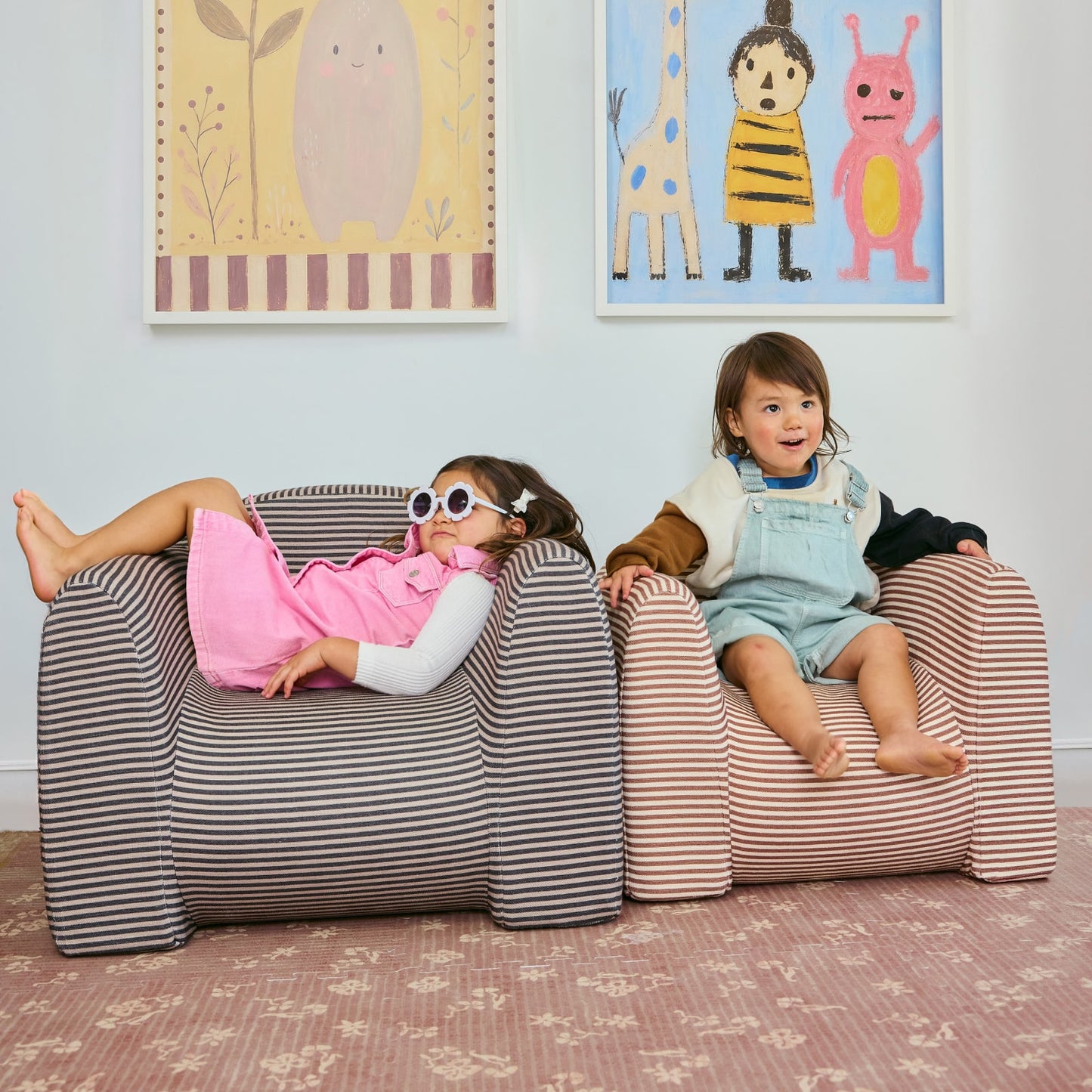Two children sitting on striped play chairs in a room with colorful wall art.