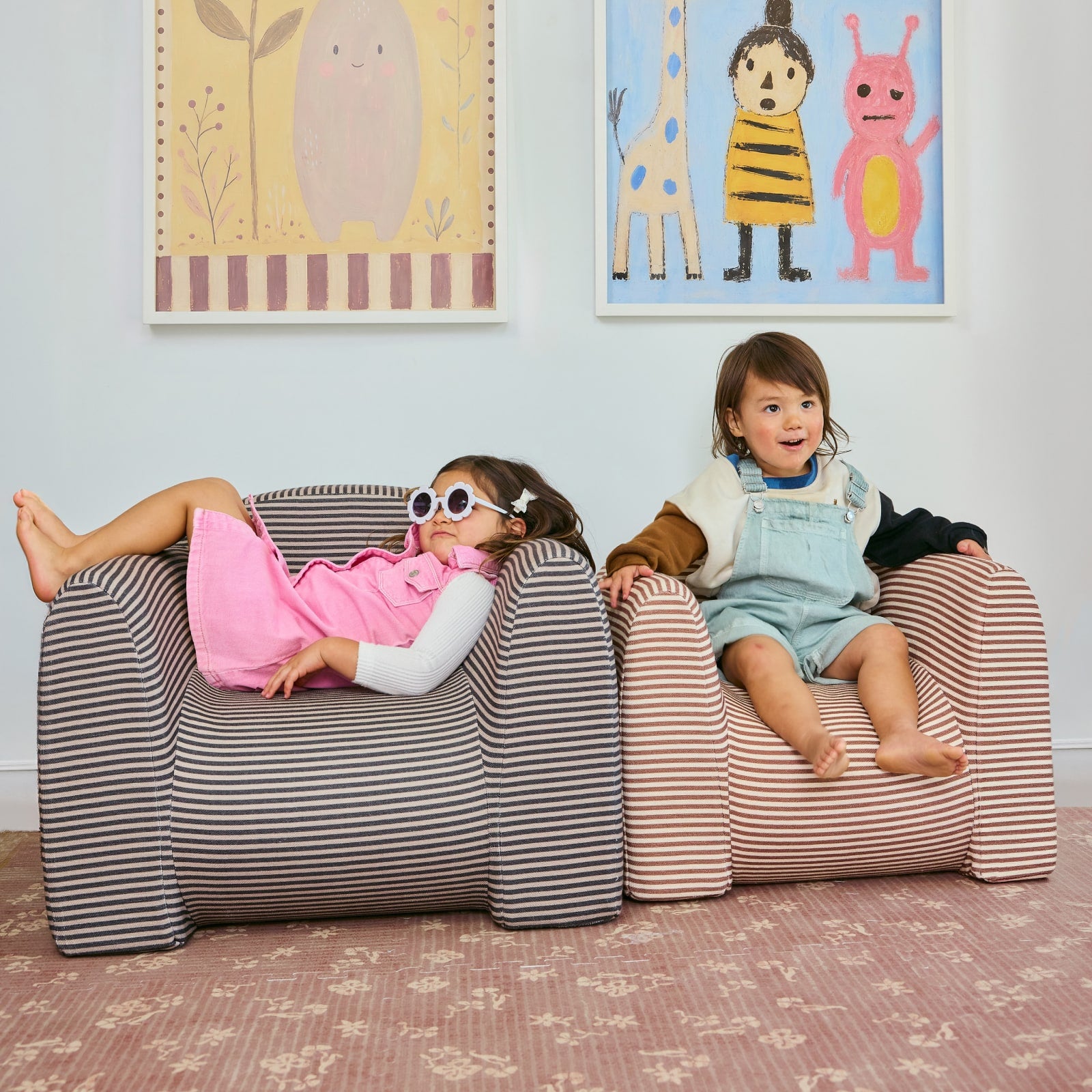 Two children sitting on striped play chairs in a room with colorful wall art.