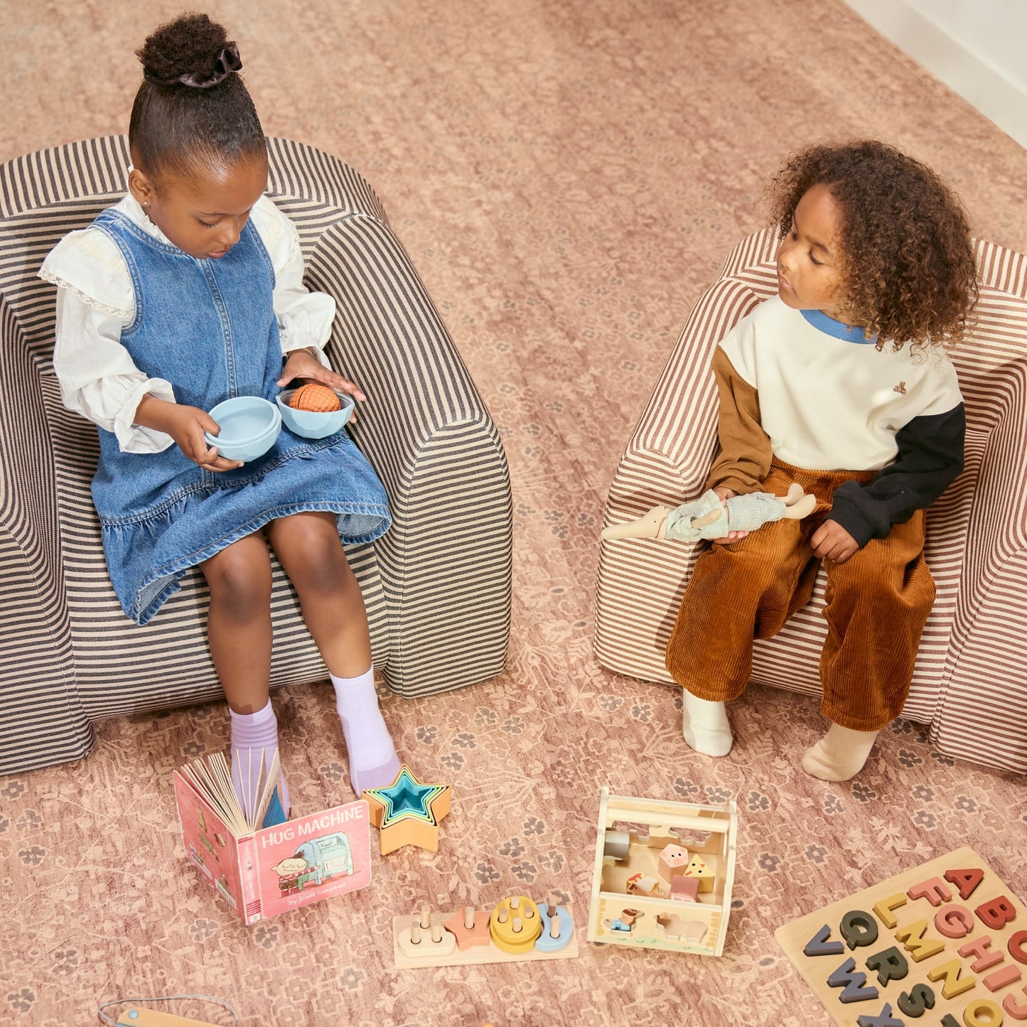 Two children sitting on patterned chairs with toys around them on a brown patern play mat.