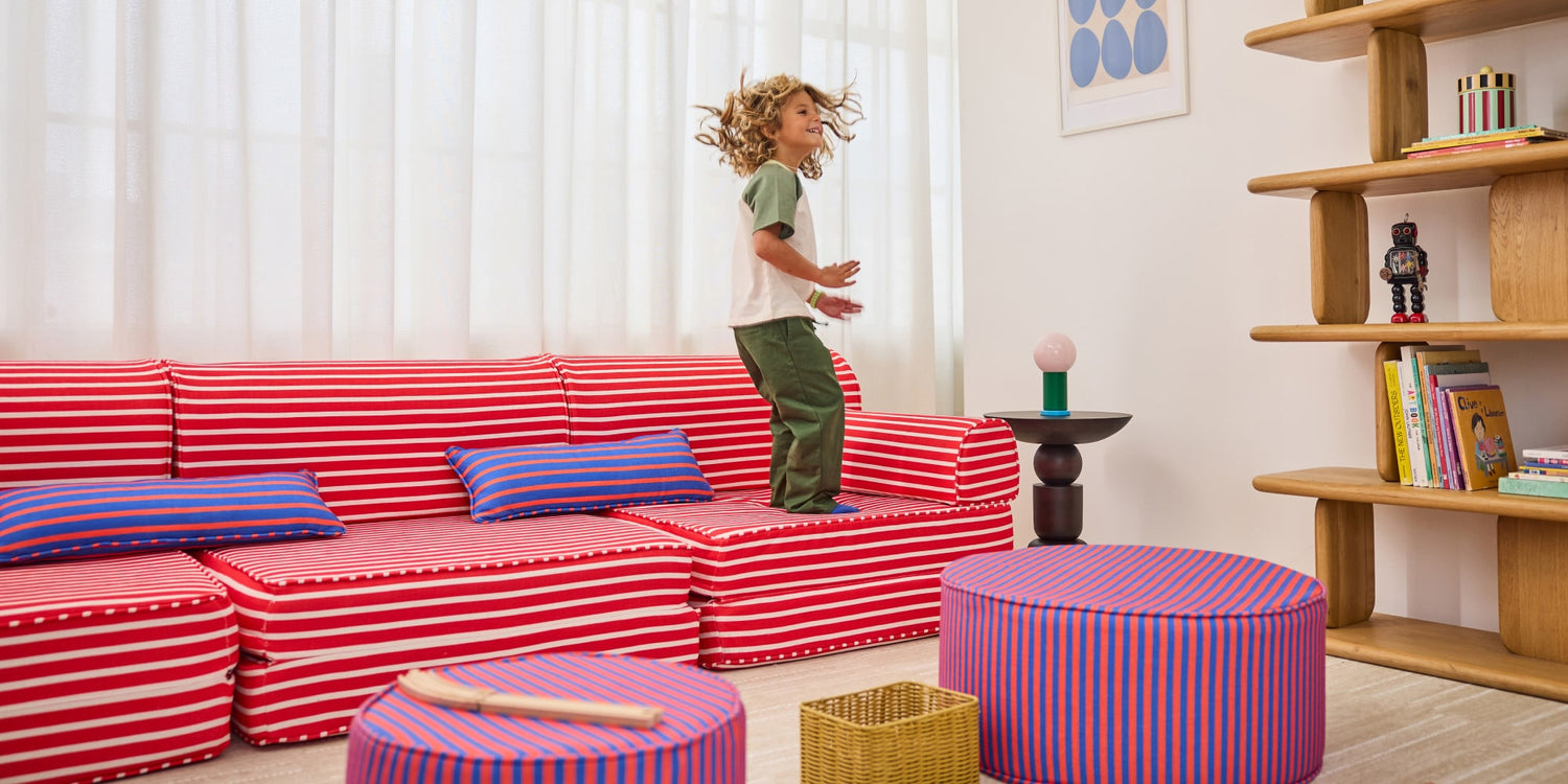 A child stands on a red and white striped sectional sofa in a bright, modern room with wooden shelves, patterned cushions, and striped ottomans.