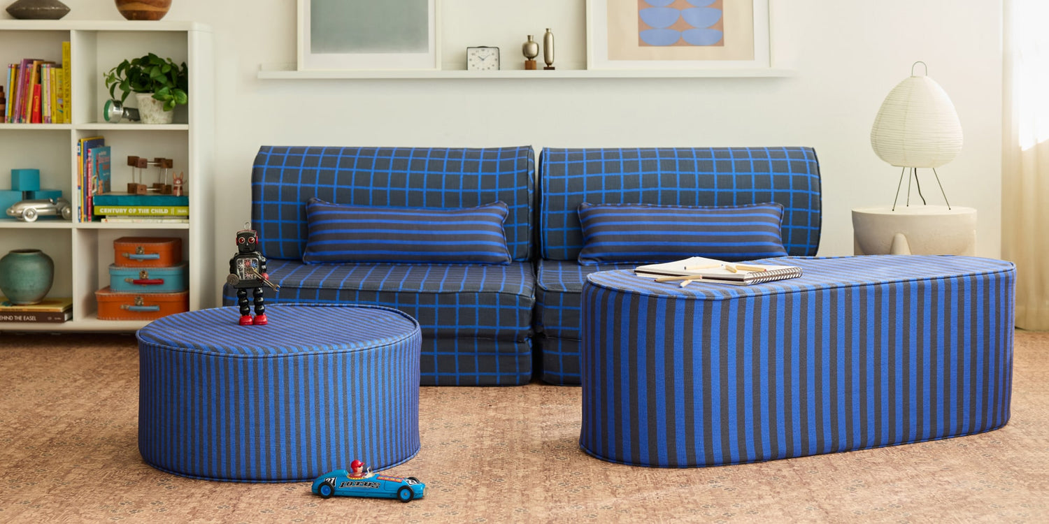 A styled living room scene featuring a blue and black plaid sofa, a matching blue striped ottoman, and a curved blue striped coffee table, set against a white wall with a bookshelf and lamp.