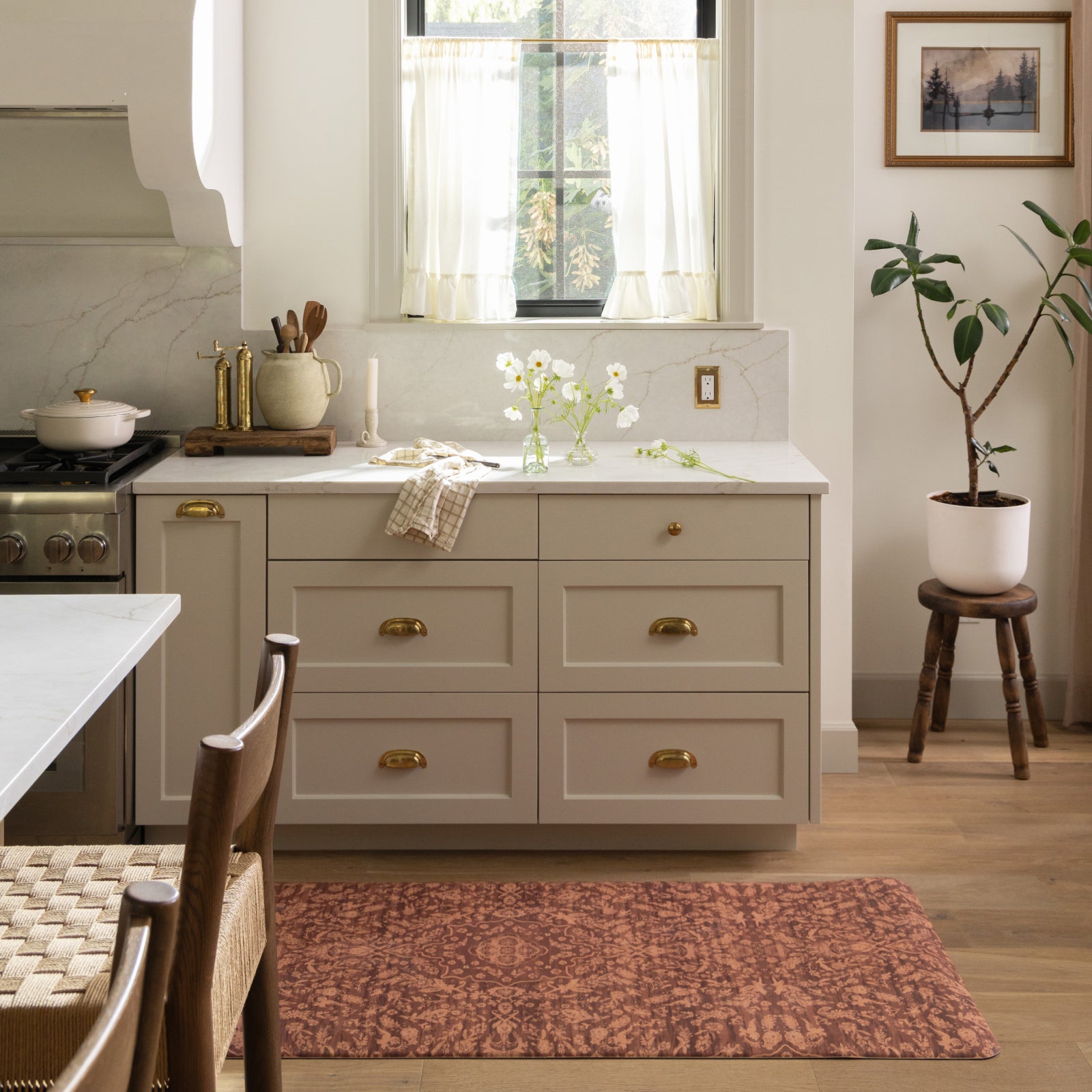 A styled kitchen interior featuring a set of six light beige cabinets with gold handles, a white marble backsplash, a patterned terracotta rug, and a potted plant on a wooden stool.