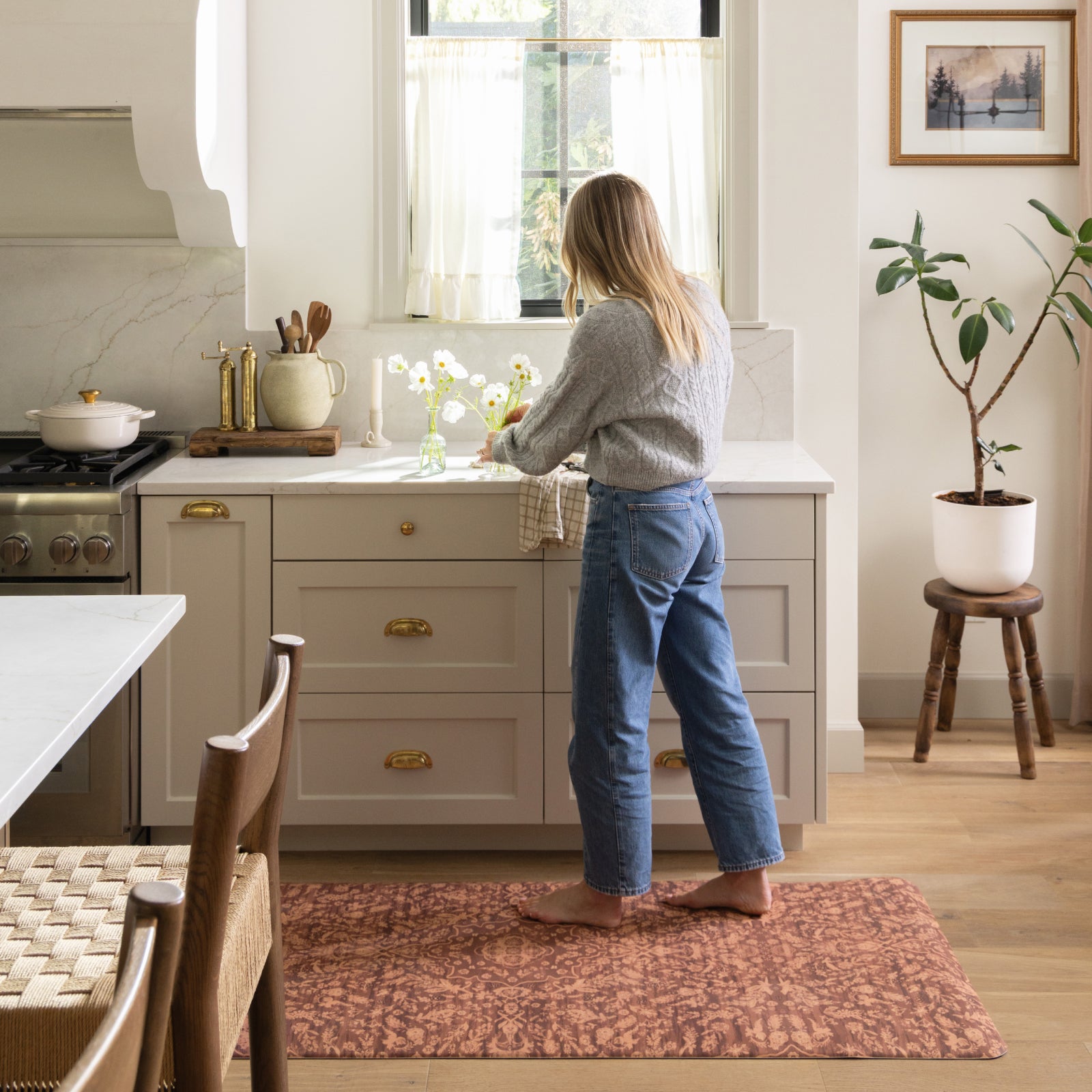A woman stands barefoot on a textured rust-brown rectangular rug in a bright kitchen, facing a white sink and light gray cabinets. The rug is placed on a light wood floor beside the counter.
