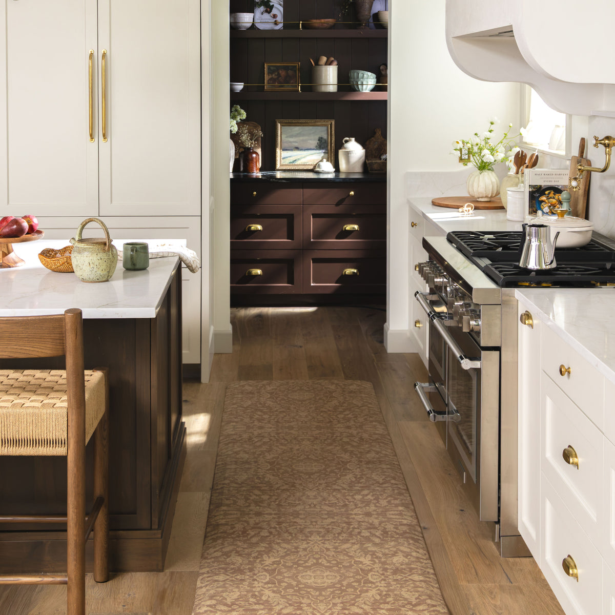 A lifestyle shot of a kitchen interior featuring a long brown patterned runner rug on a light wood floor, flanked by white cabinets, a dark wood island, and a stainless steel gas range.