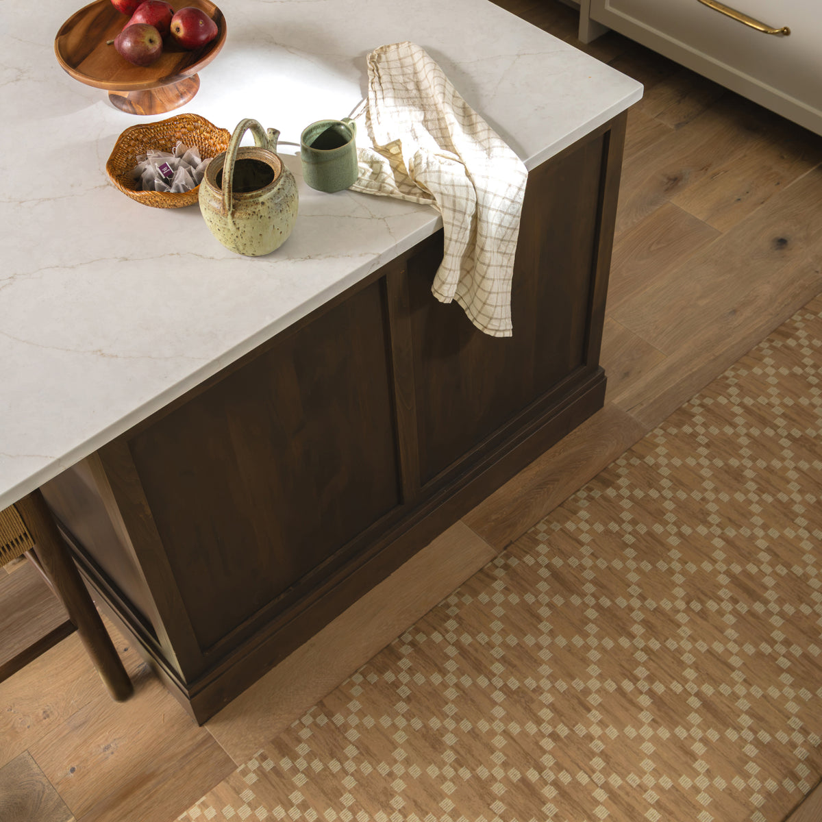 A kitchen island with a white marble-look countertop and dark brown wood cabinet base, styled with ceramic mugs, a checkered cloth, and fruit bowls, set on a wood floor with a beige patterned rug.
