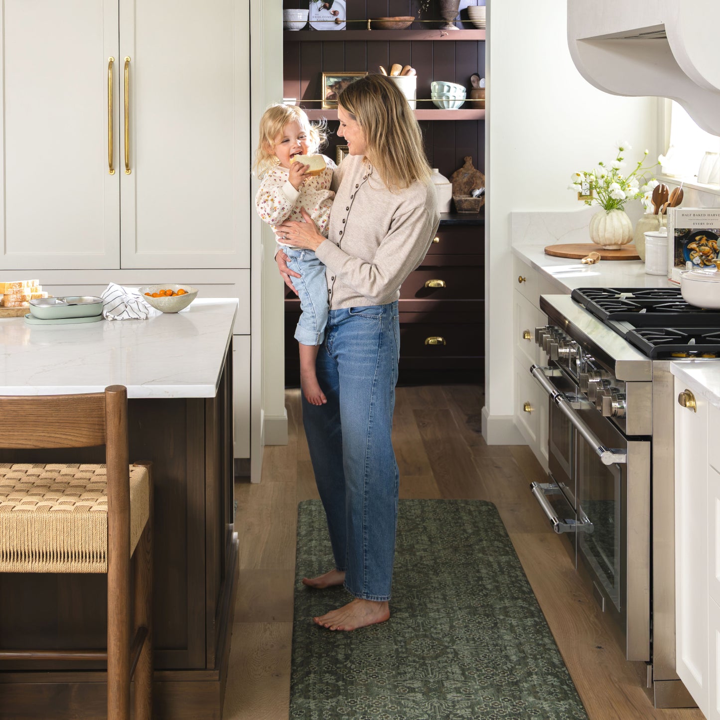 A woman in a beige top and blue jeans holds a toddler in a bright, modern kitchen. The toddler wears a cream patterned shirt and light blue jeans.