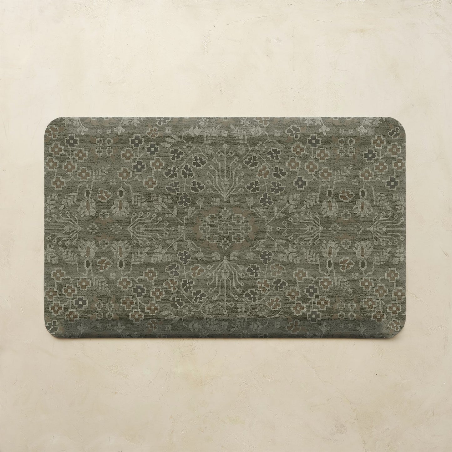 A rectangular floor mat with rounded corners featuring an intricate distressed floral and medallion pattern in muted gray-green and beige tones, displayed against a textured beige wall.