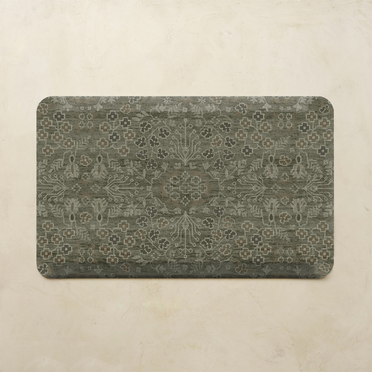 A rectangular floor mat with rounded corners featuring an intricate distressed floral and medallion pattern in muted gray-green and beige tones, displayed against a textured beige wall.