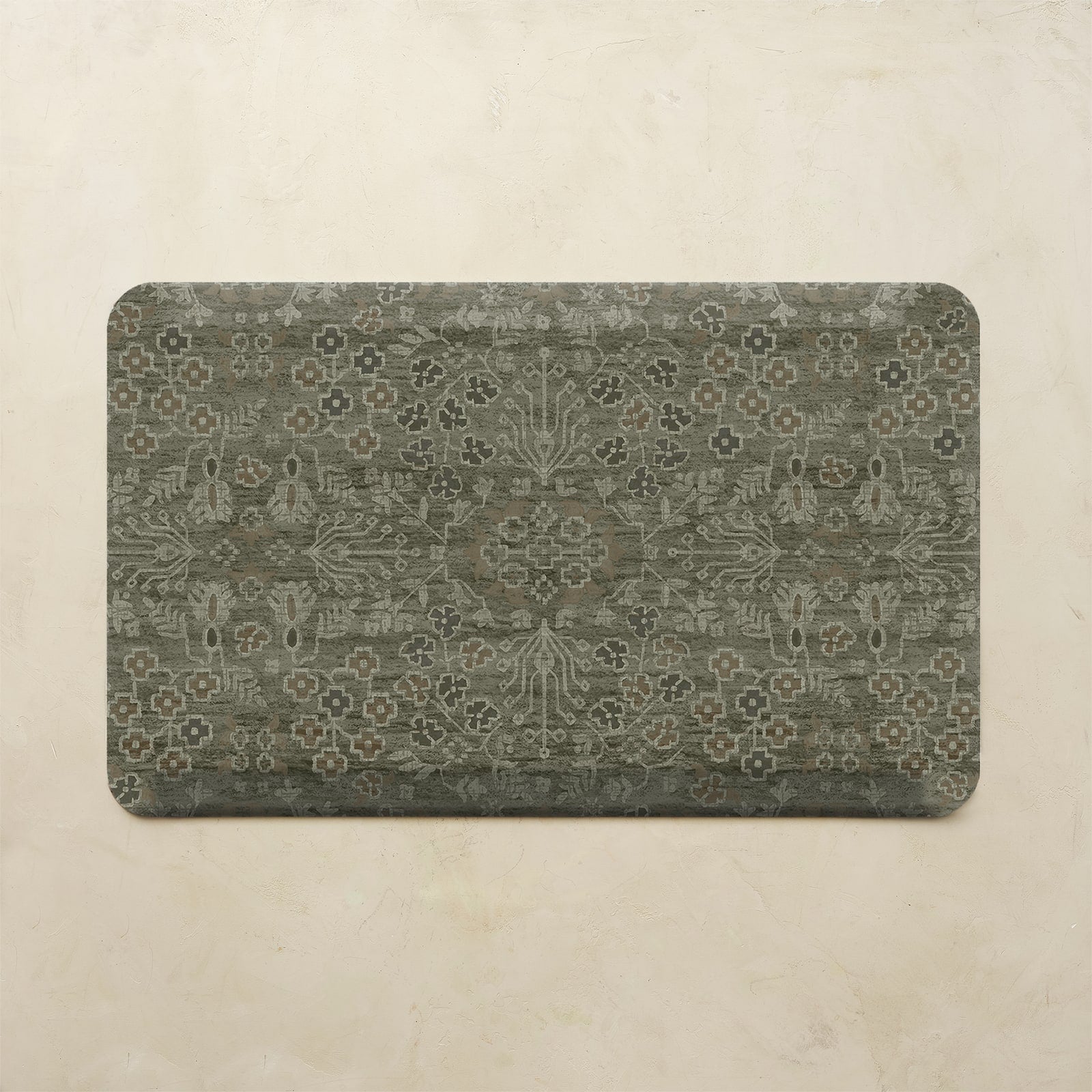 A rectangular floor mat with rounded corners featuring an intricate distressed floral and medallion pattern in muted gray-green and beige tones, displayed against a textured beige wall.