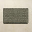 A rectangular floor mat with rounded corners featuring an intricate distressed floral and medallion pattern in muted gray-green and beige tones, displayed against a textured beige wall.