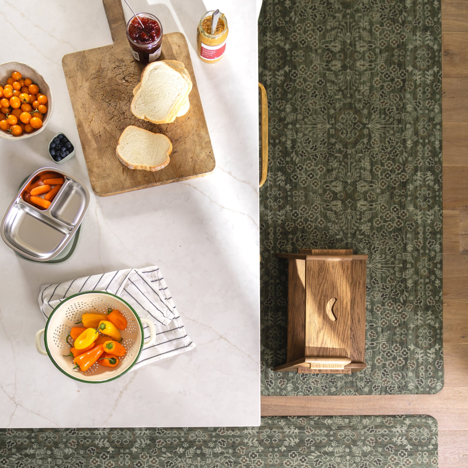 A styled overhead kitchen scene showing a dark green patterned runner rug laid along the right side of a white marble countertop, with breakfast items like bread, jam, carrots, and a wooden cutting board nearby.
