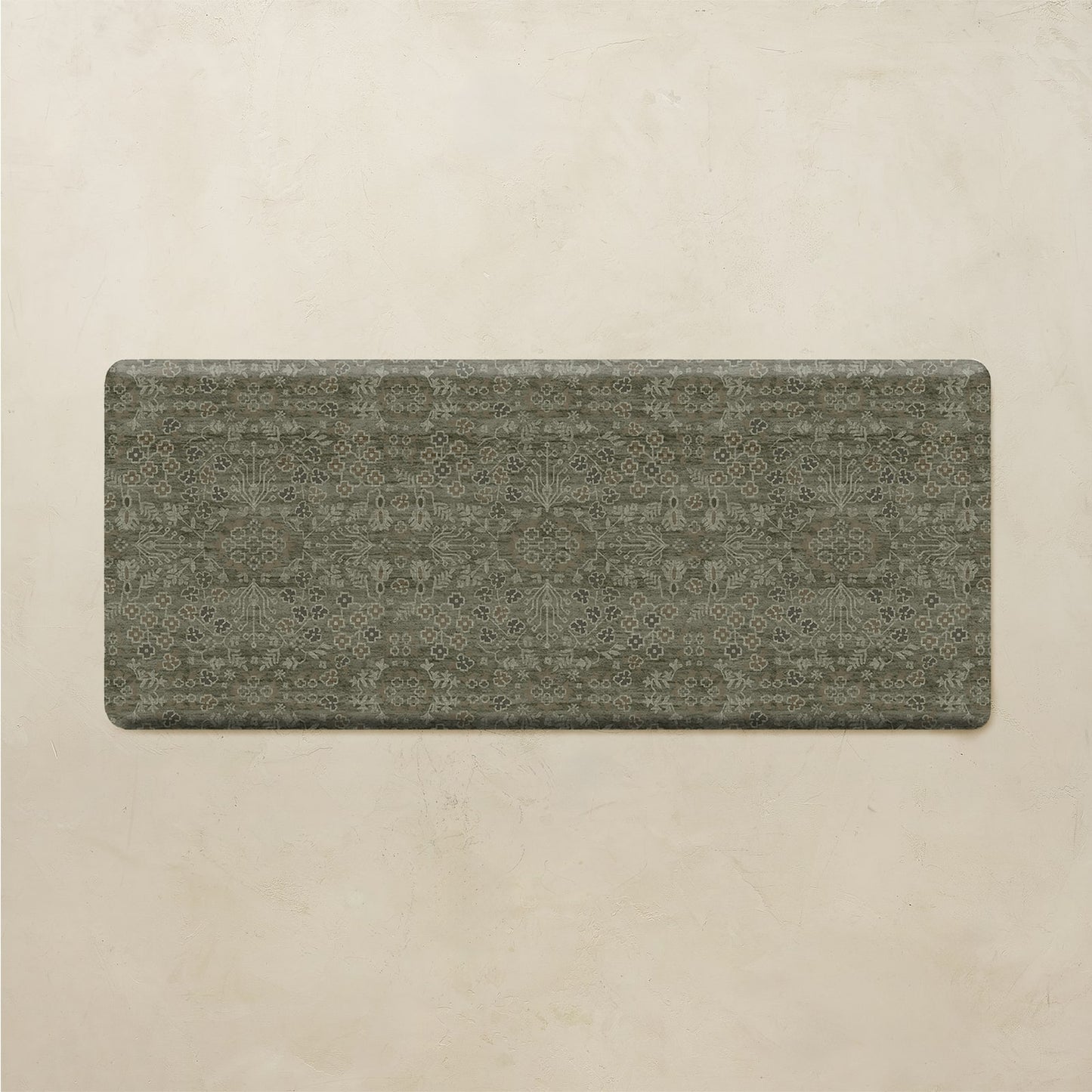 A rectangular floor mat with rounded corners, featuring a distressed, vintage-style ornamental pattern in muted olive green and gray tones, displayed against a textured beige background.