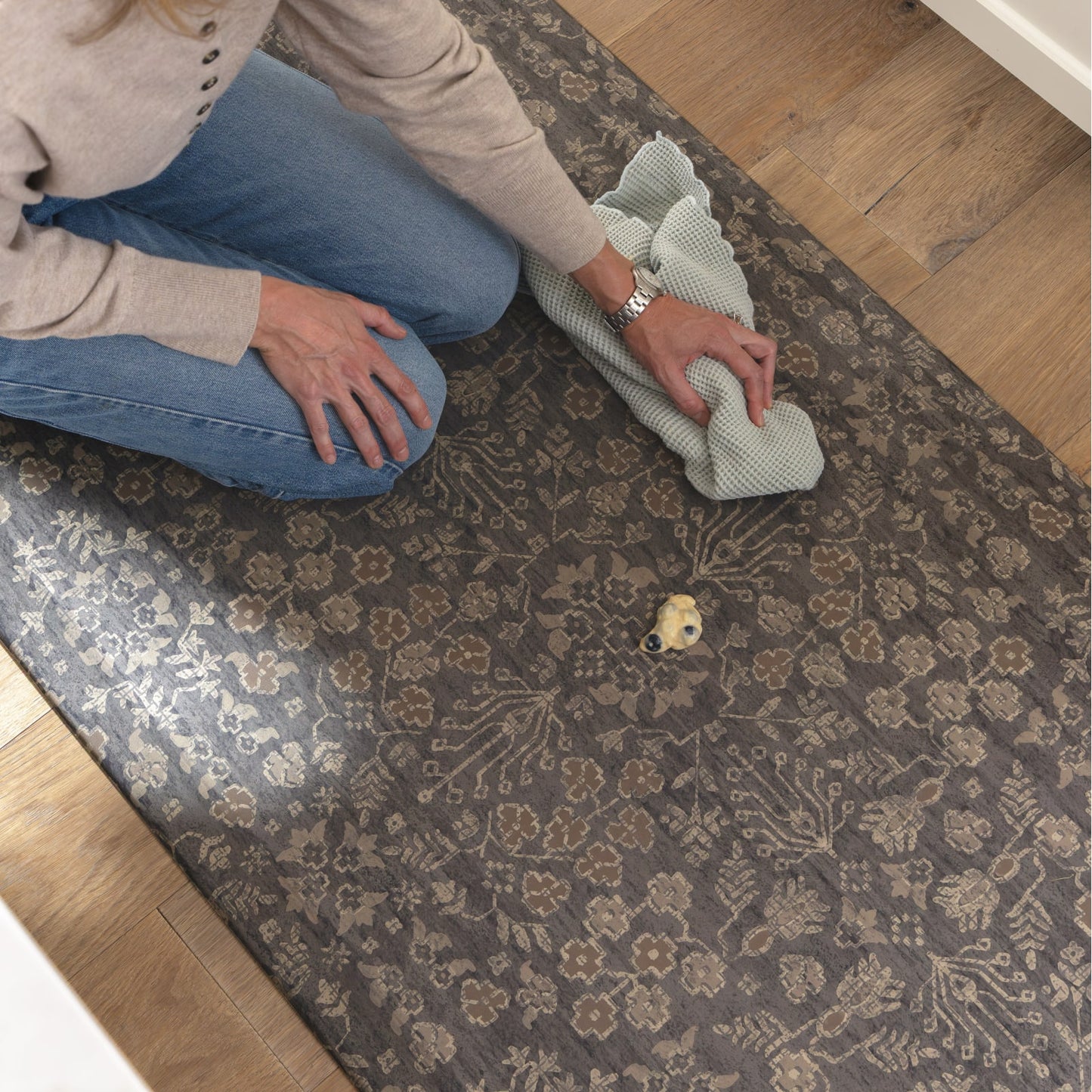 A person kneels on a brown and beige floral patterned area rug while cleaning it with a light green cloth, set on a light wood floor.