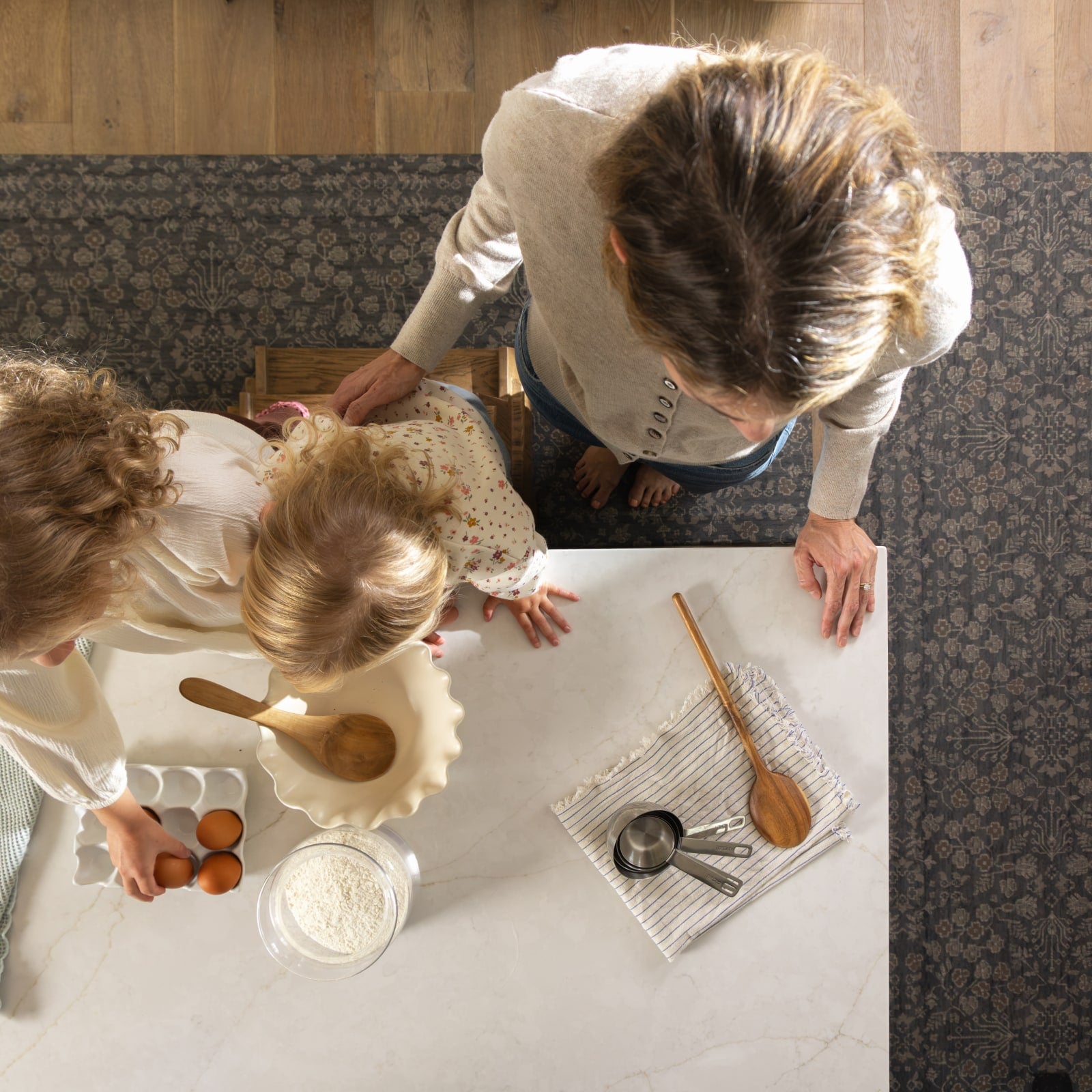 Overhead view of a woman and two children gathered around a white marble kitchen table, preparing food with a bowl, eggs, flour, measuring cups, and wooden utensils on a patterned rug.