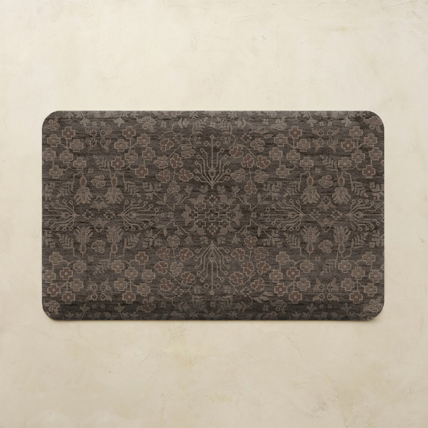 A rectangular kitchen mat with rounded corners featuring an intricate, faded damask-style floral pattern in dark brown and charcoal gray, displayed on a textured beige background.