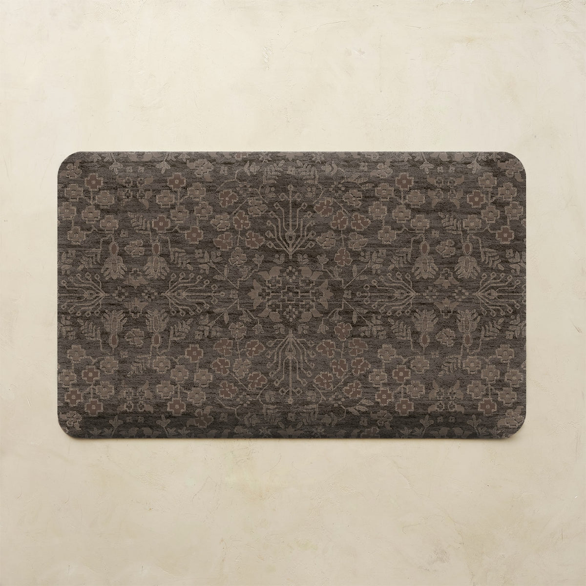 A rectangular kitchen mat with rounded corners featuring an intricate, faded damask-style floral pattern in dark brown and charcoal gray, displayed on a textured beige background.