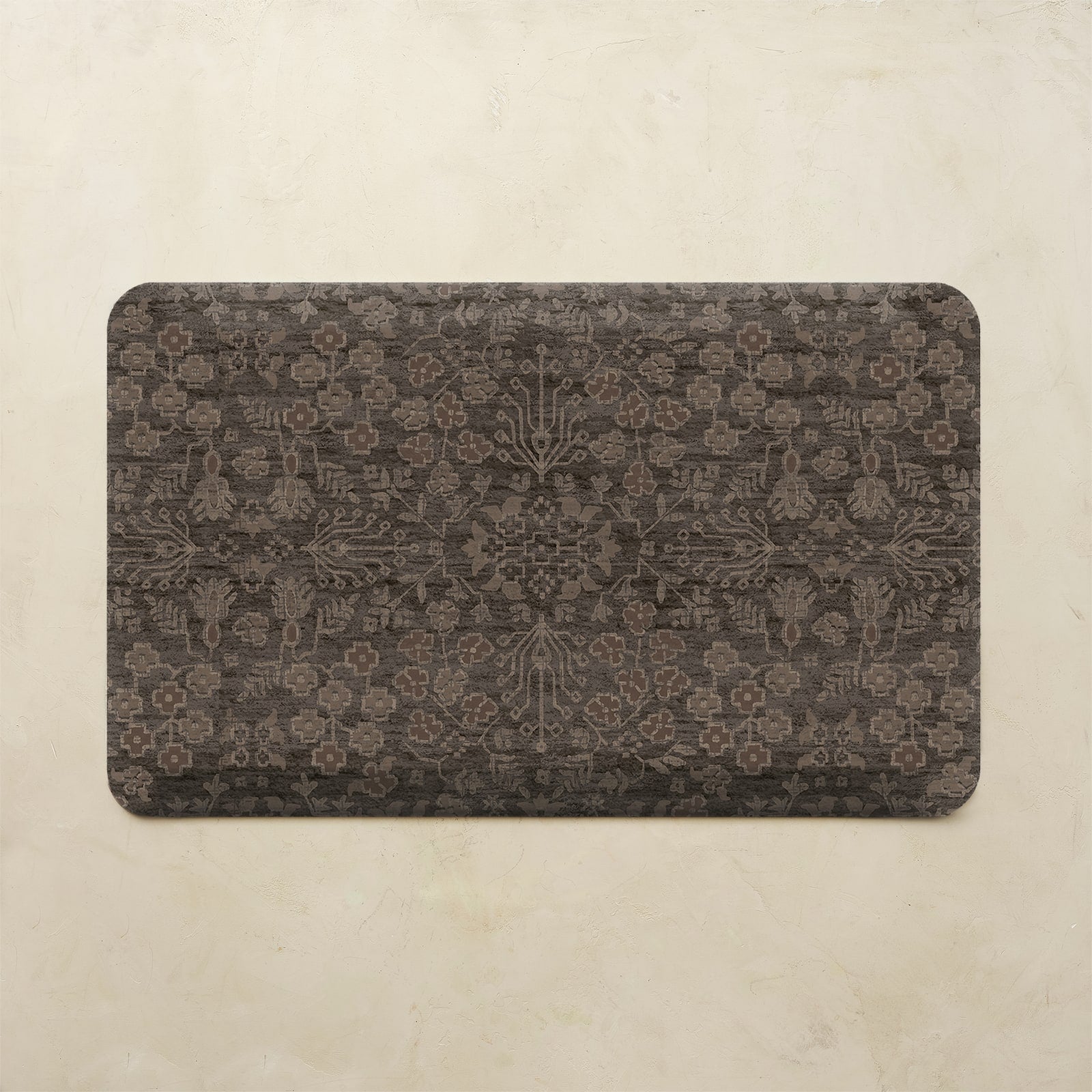 A rectangular kitchen mat with rounded corners featuring an intricate, faded damask-style floral pattern in dark brown and charcoal gray, displayed on a textured beige background.