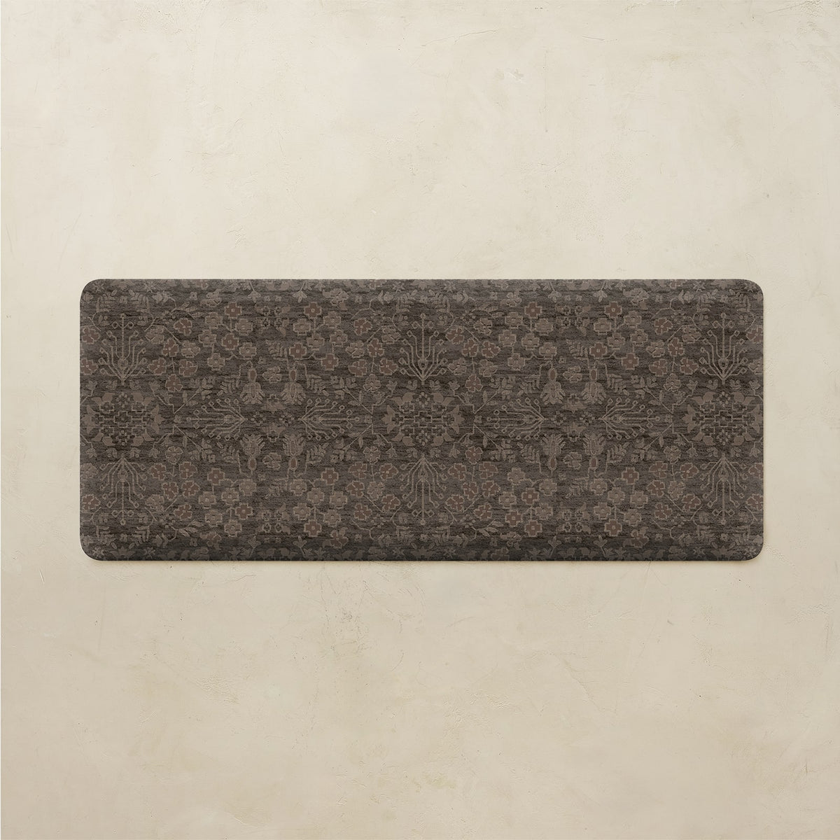 A rectangular floor mat with rounded corners, featuring an intricate dark brown and charcoal gray traditional pattern, displayed against a textured beige background.