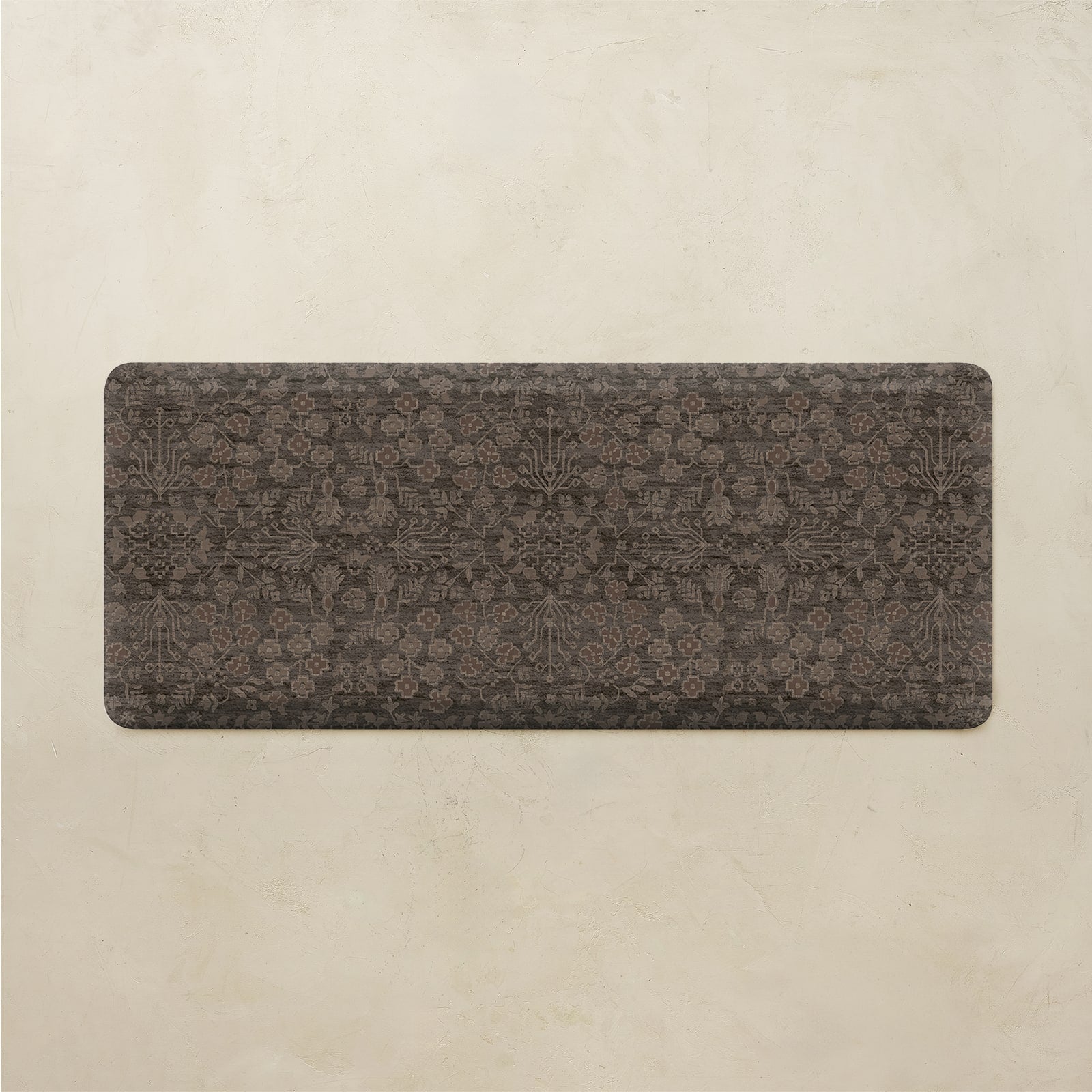 A rectangular floor mat with rounded corners, featuring an intricate dark brown and charcoal gray traditional pattern, displayed against a textured beige background.