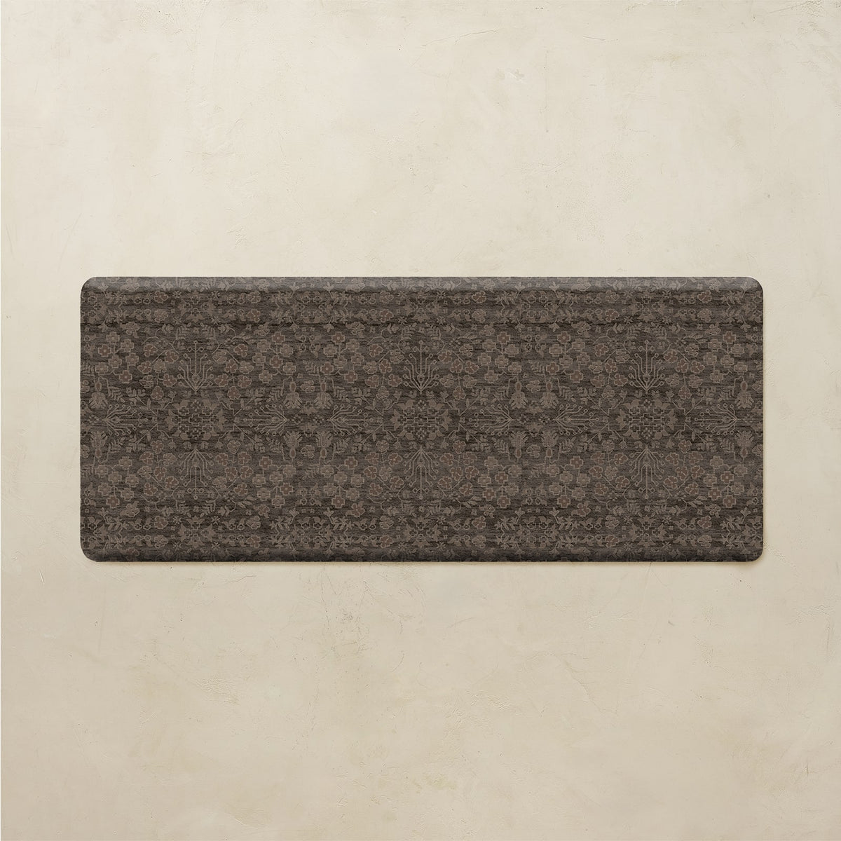 A rectangular floor mat with rounded corners, featuring a dark brown and charcoal gray traditional floral pattern, displayed against a textured beige background.