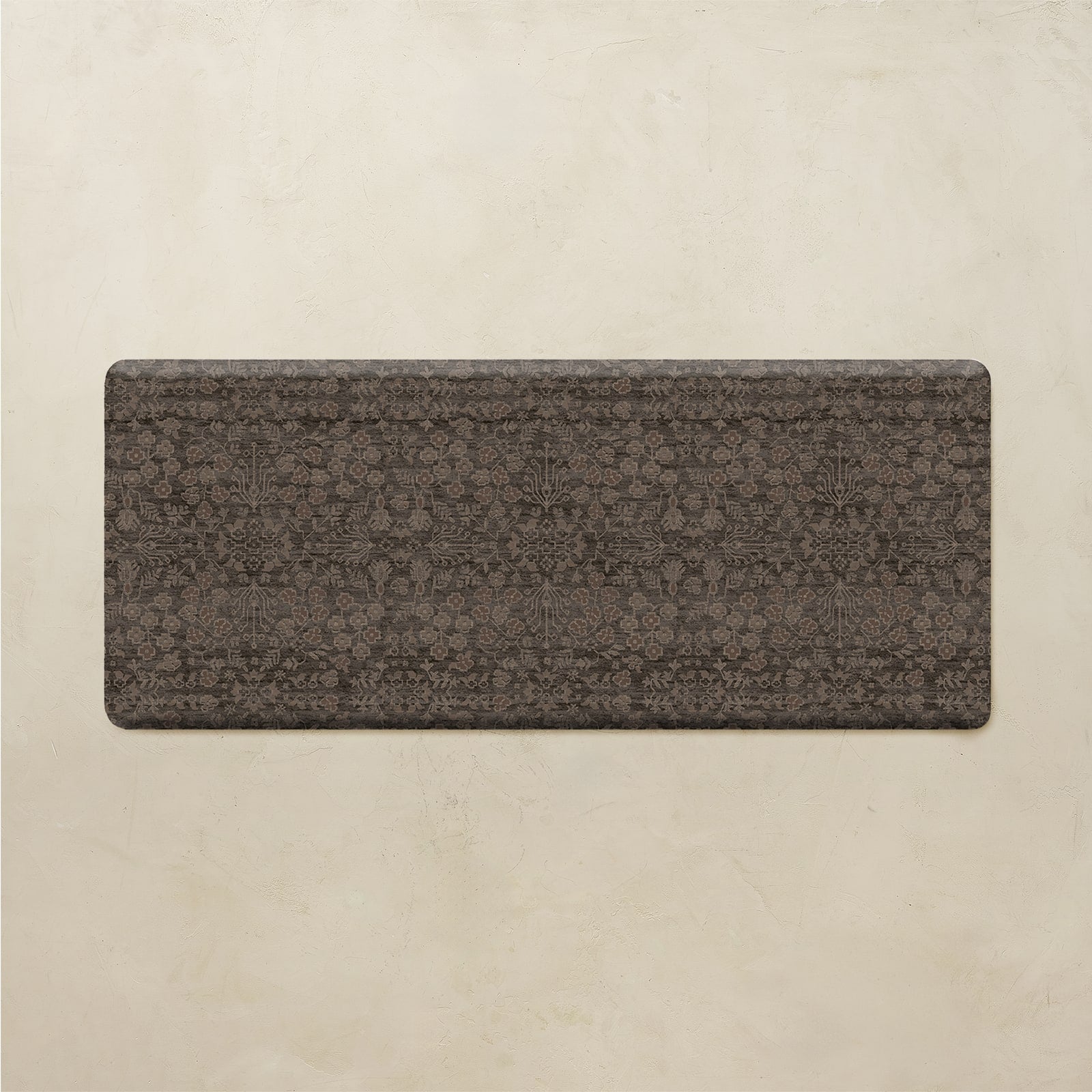 A rectangular floor mat with rounded corners, featuring a dark brown and charcoal gray traditional floral pattern, displayed against a textured beige background.