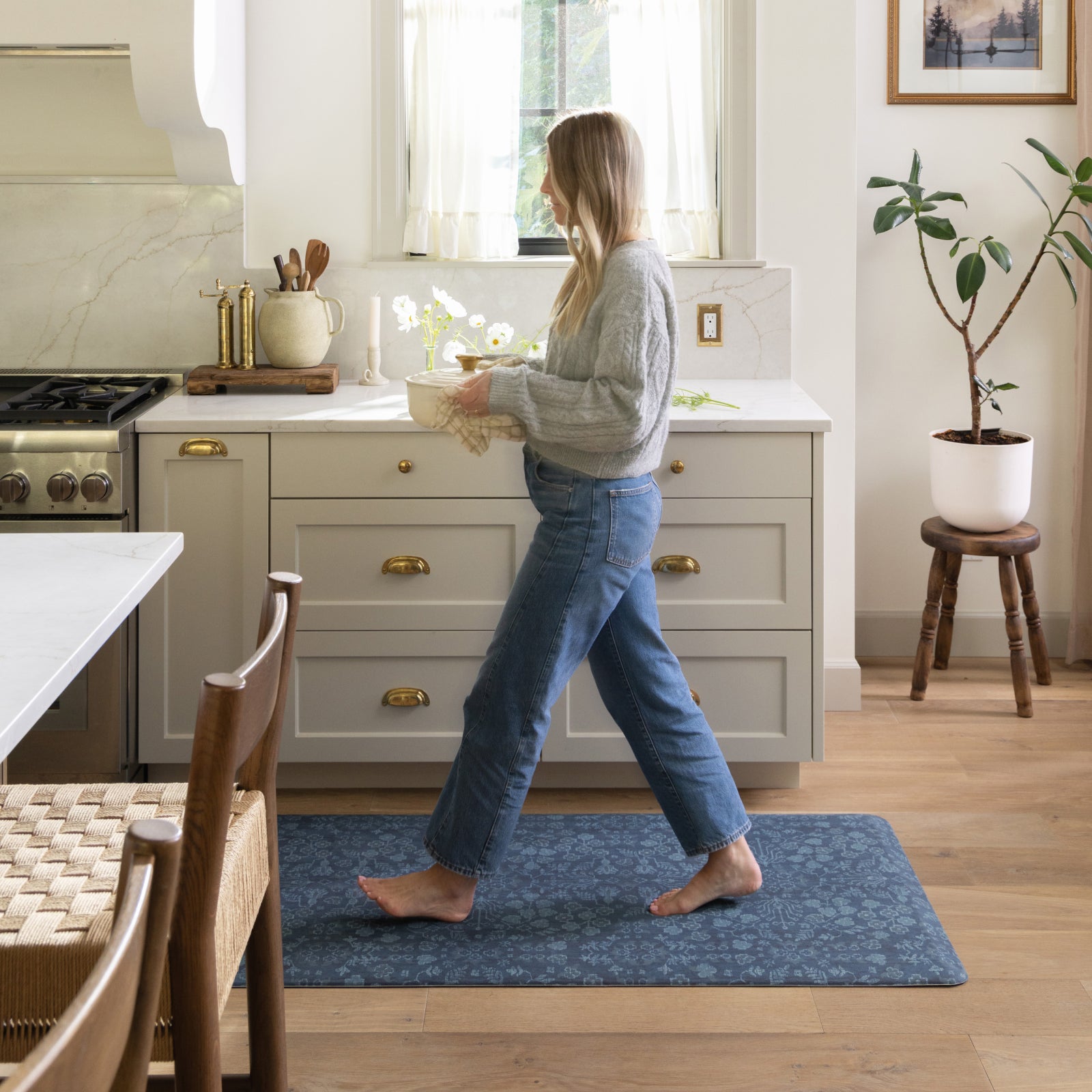 A woman walks barefoot across a blue patterned area rug in a bright, modern kitchen with light gray cabinets and wood floors.