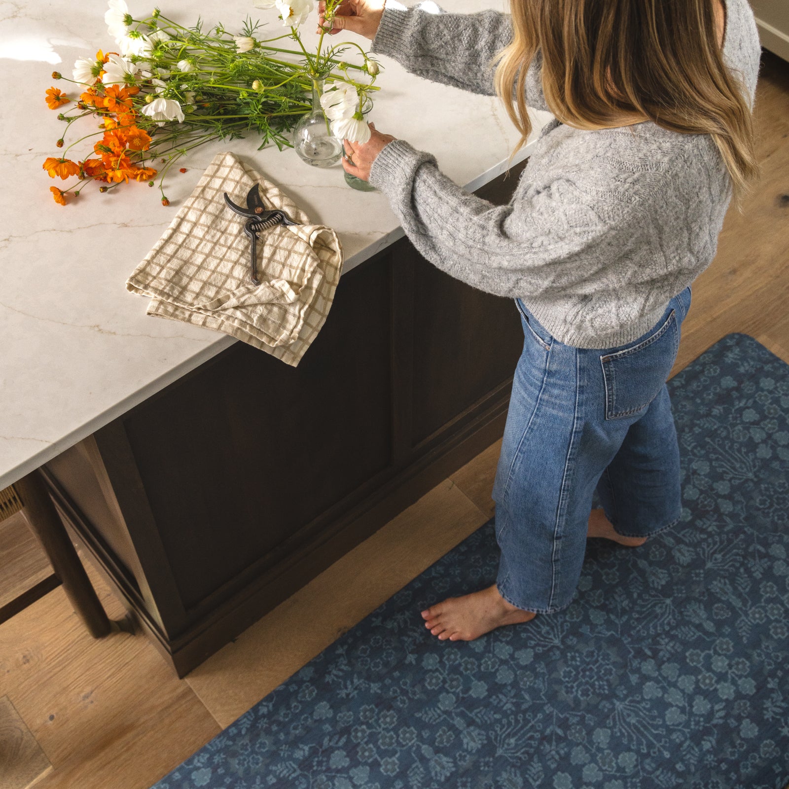 A woman in a gray sweater and blue jeans stands barefoot on a blue patterned rug beside a kitchen island, arranging white flowers in a glass vase. The scene includes orange flowers on the marble countertop and a folded beige checkered cloth with a black-handled tool.