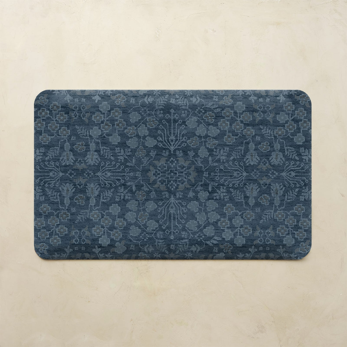 A rectangular bath mat with rounded corners featuring a dark navy blue and slate gray traditional floral pattern, displayed on a textured beige stone surface.