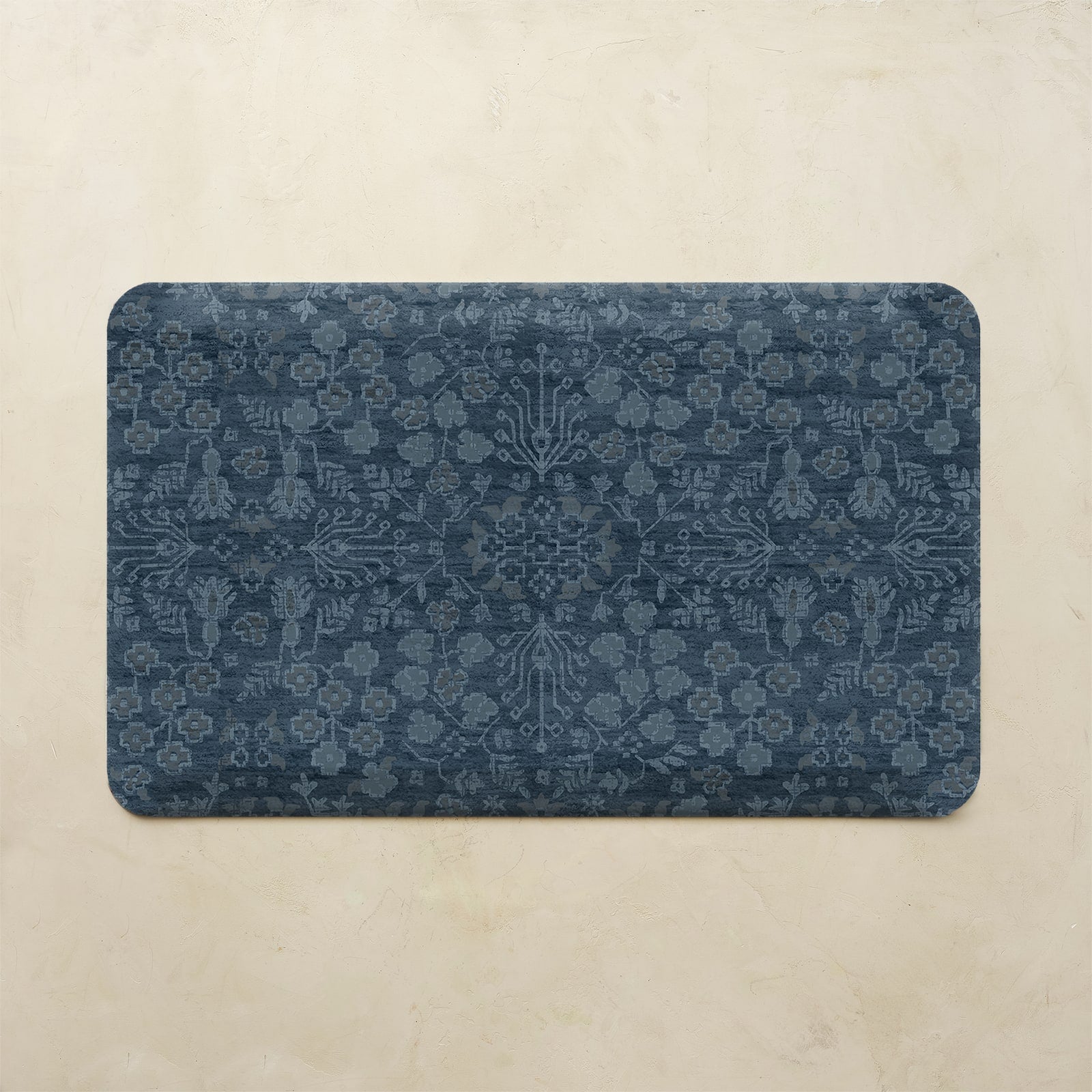 A rectangular bath mat with rounded corners featuring a dark navy blue and slate gray traditional floral pattern, displayed on a textured beige stone surface.