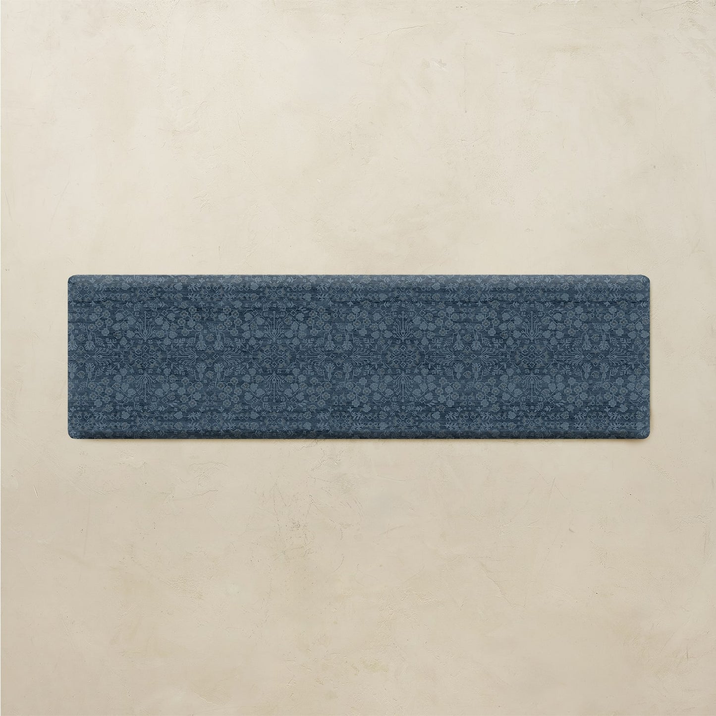 A rectangular blue textured runner rug with a subtle floral pattern, displayed horizontally against a warm beige textured wall background.
